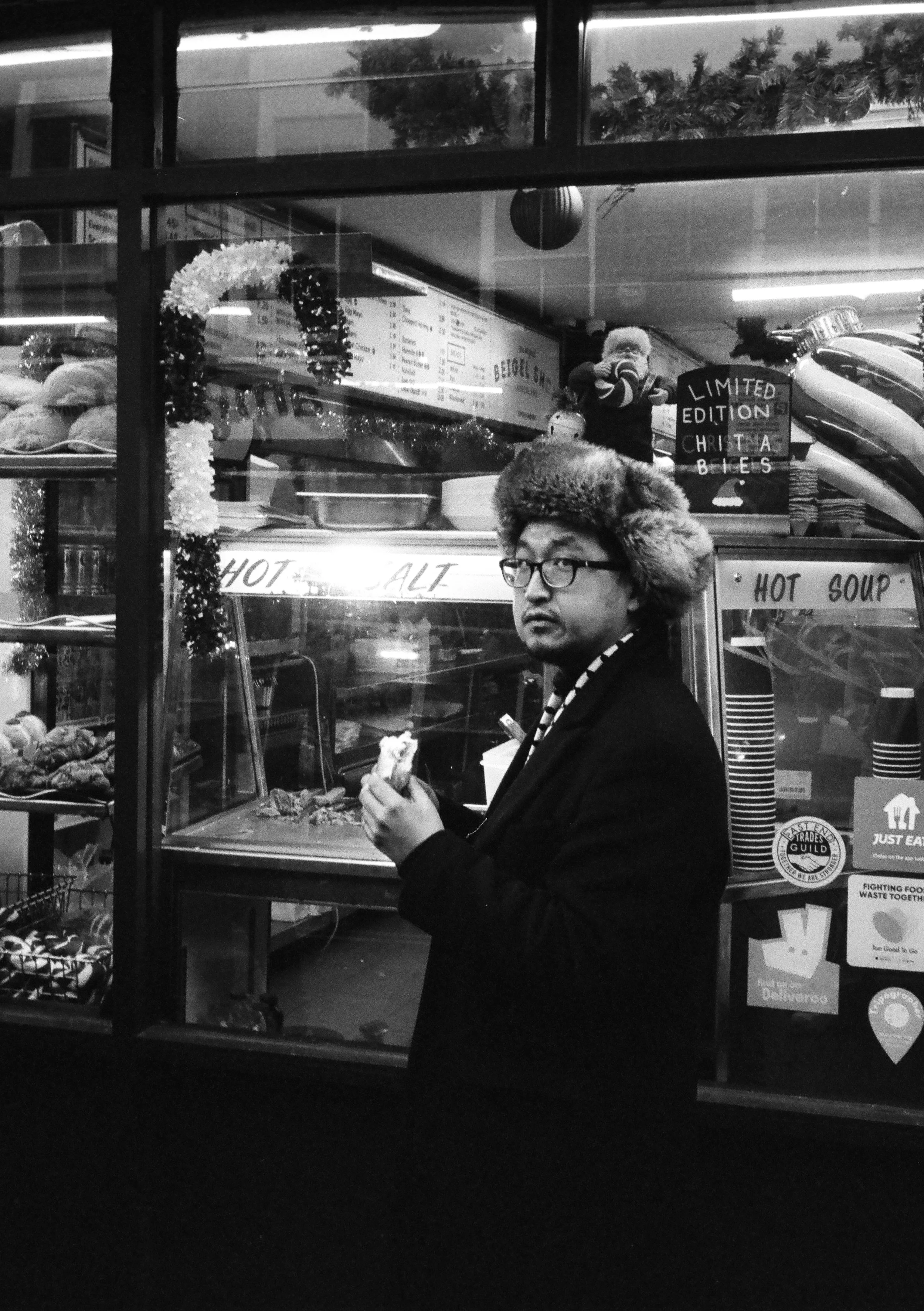 A man wearing glasses, a black coat, and a furry hat standing outside a food stall, holding food in his hand, with various signs and decorations in the background.
