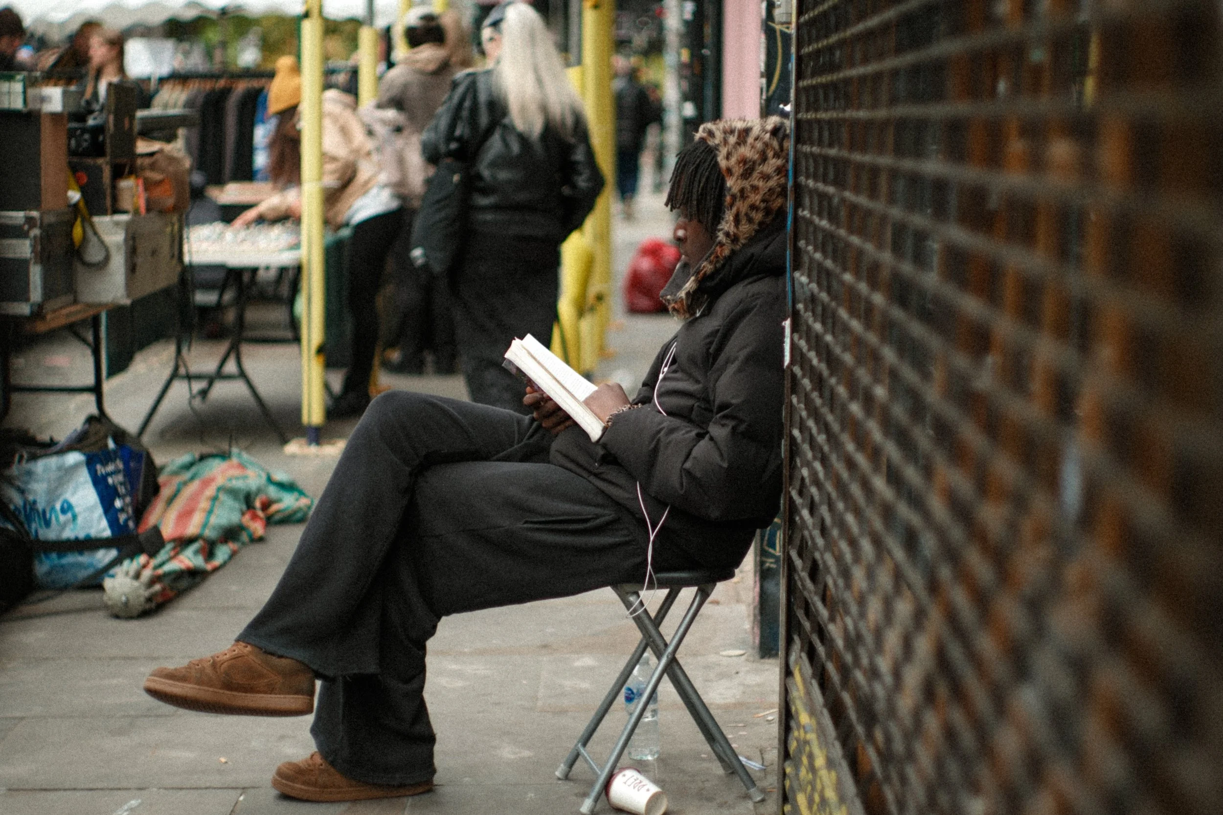 A person with a leopard print hoodie under a black jacket, reading a book while sitting on a folding chair on a city sidewalk, with a water bottle and coffee cup on the ground nearby, and a crowded outdoor market in the background.