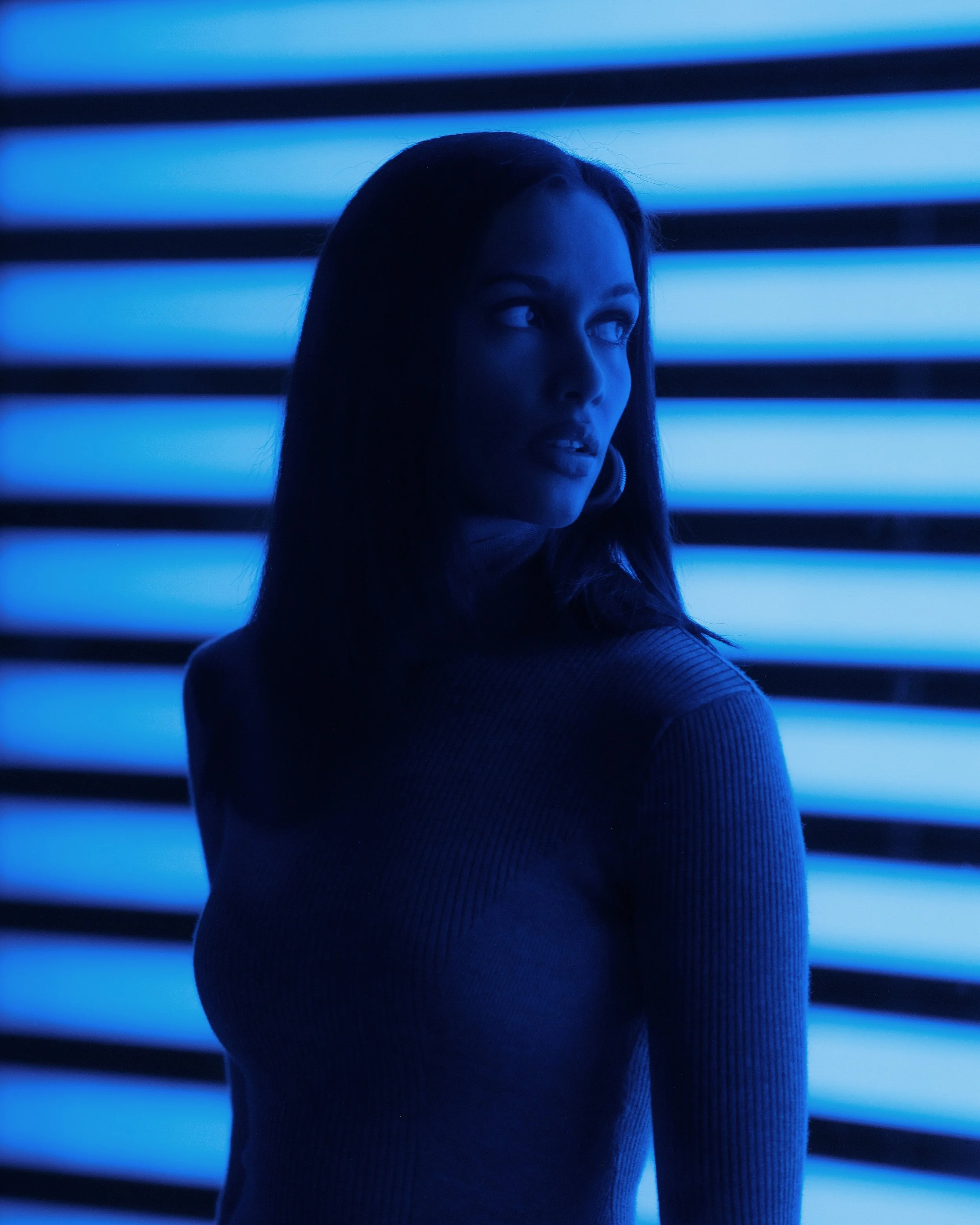 A woman with dark hair looking over her shoulder in front of illuminated horizontal blue and black stripes.