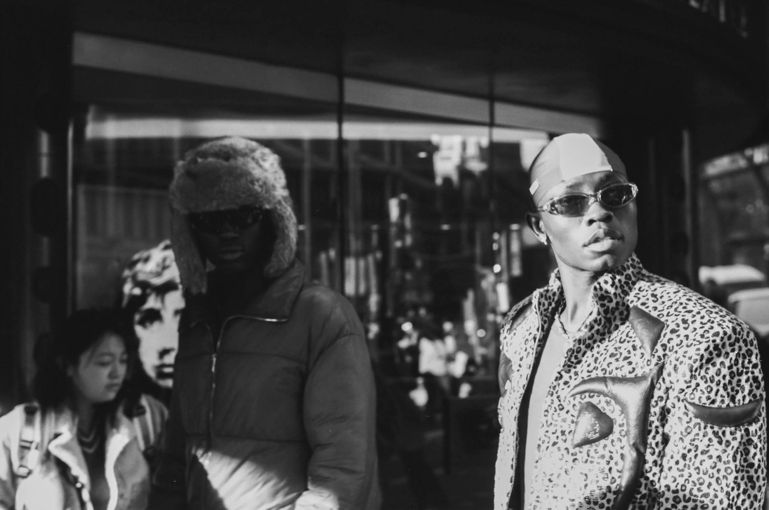 Two women standing outdoors, one with dark skin wearing sunglasses, a shiny patterned jacket, a head covering, and an expression of confidence; the other with dark skin wearing a jacket with a fur collar and arriving at his face with sunglasses, the 