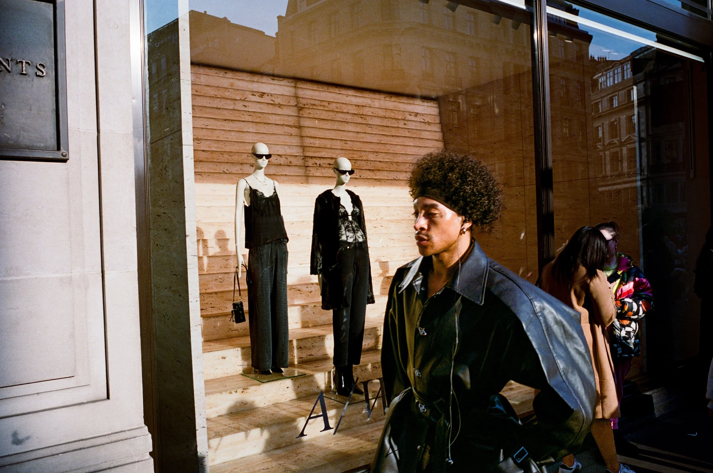 City street scene with mannequins in storefront window display dressed in black clothing, and a young man with curly hair and leather jacket walking past.