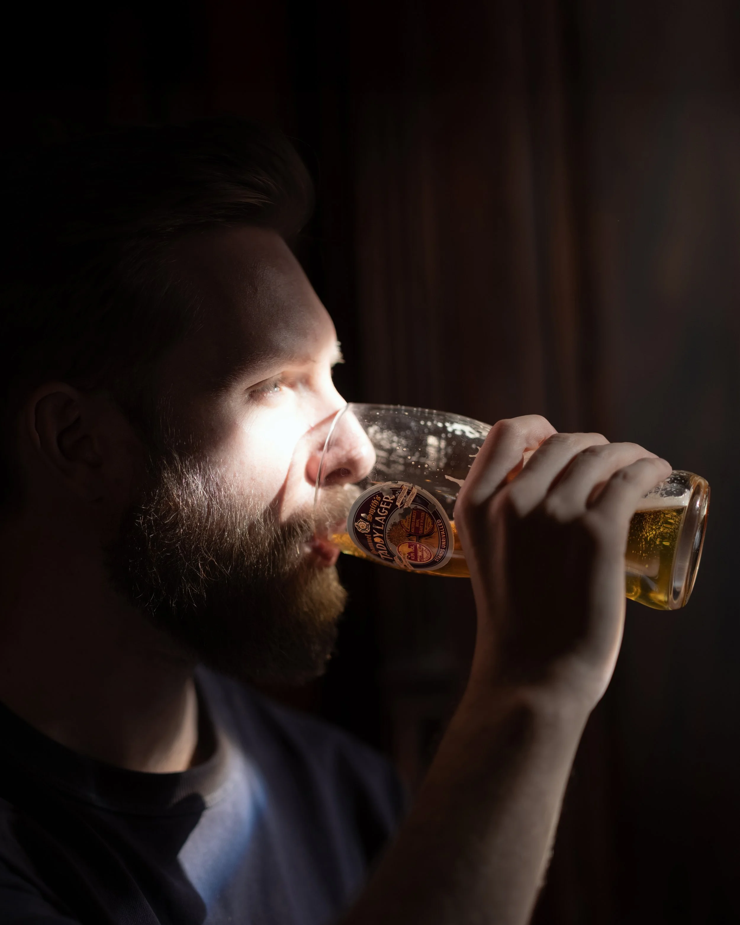 A man with a beard drinking beer from a glass in a dimly lit room.