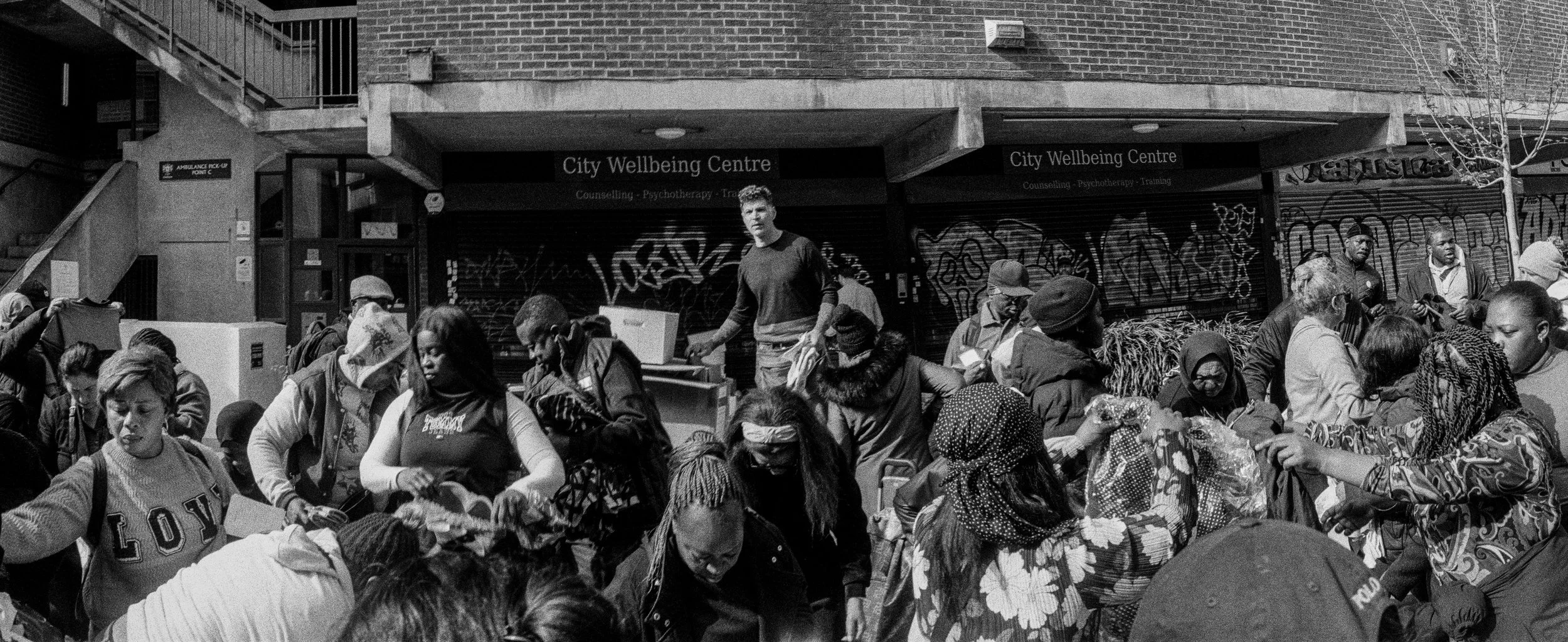 A black and white photo of a crowd of people queued outdoors in front of a building labeled 'City Wellbeing Centre' with Graffiti on the shutters, and a sign indicating 'Ambulance pick-up point' on the left side.