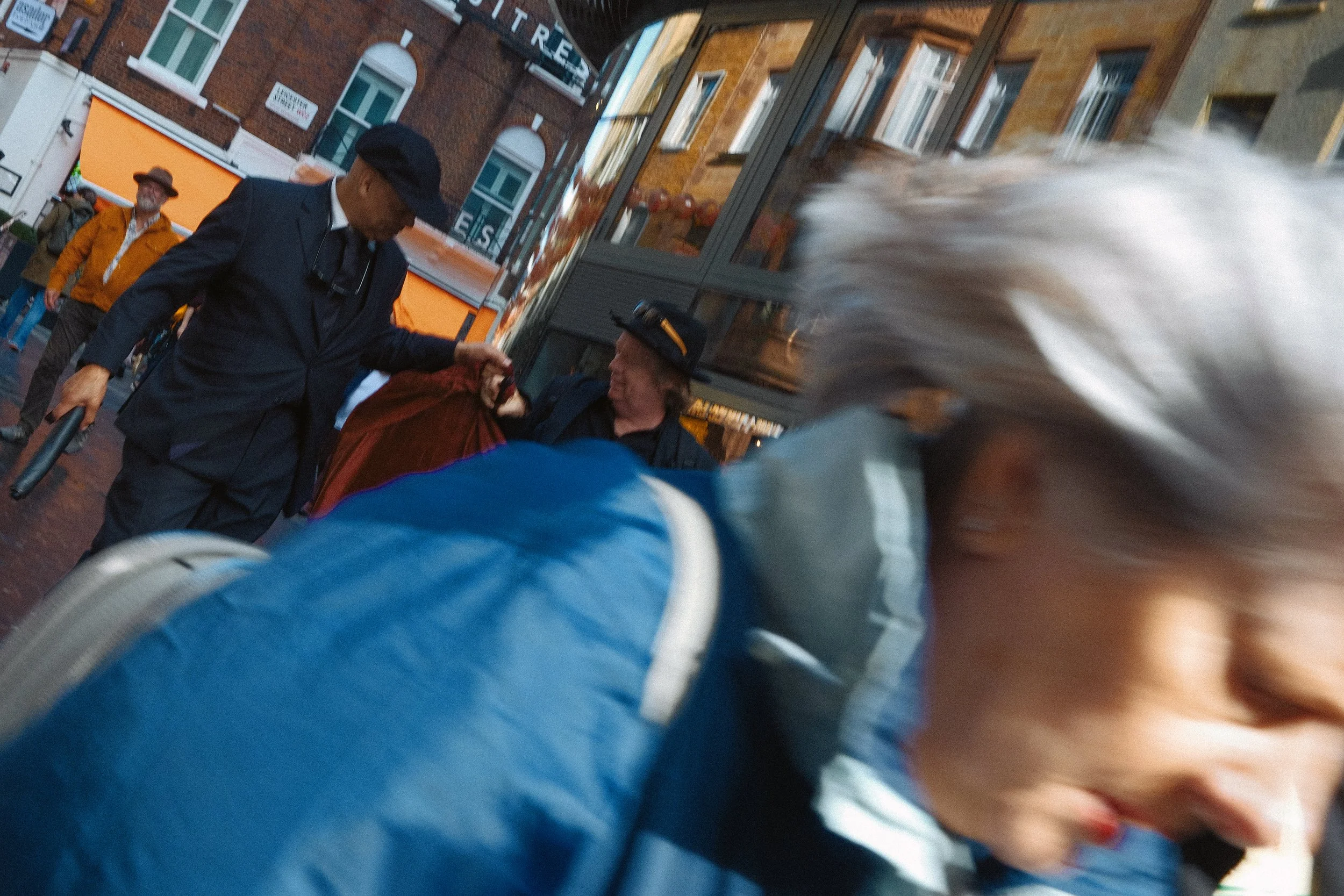 Blurred close-up of a person with gray hair and glasses, with a background showing two men in dark suits and hats engaging with a red object, on a city street with brick buildings and pedestrians.