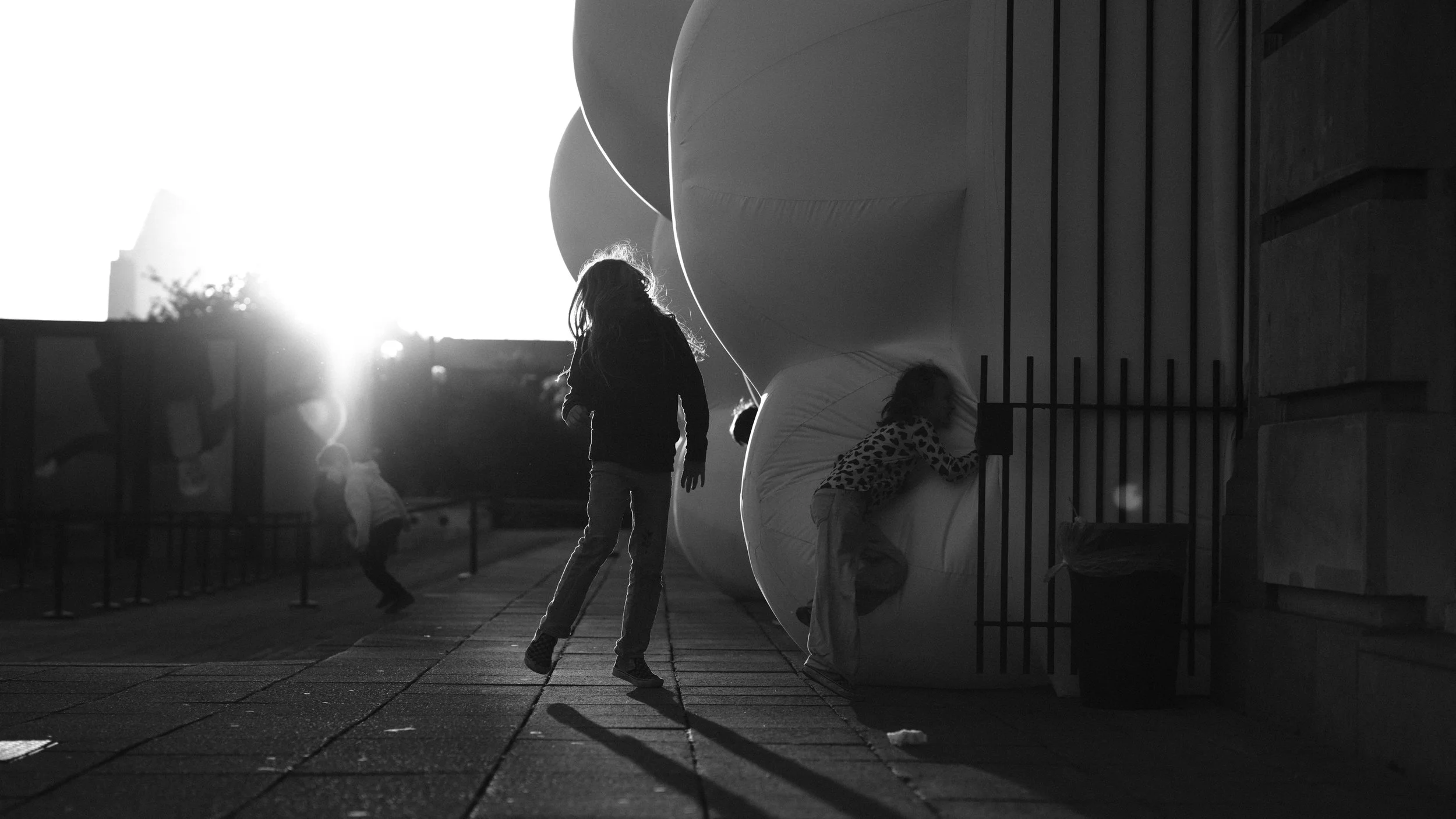 Children playing outside during sunset with some exploring a large inflatable structure and others running, in black and white.