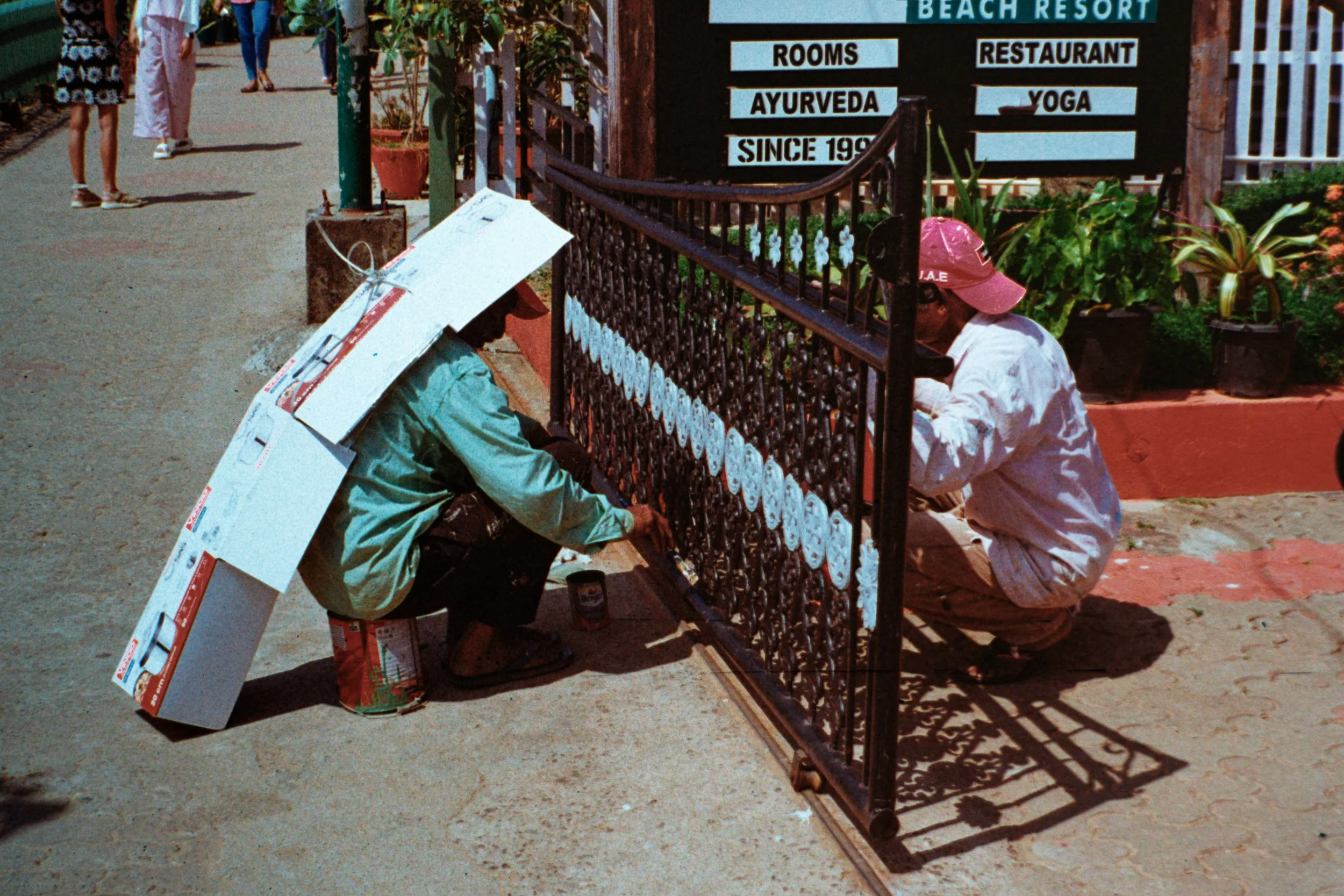 Two men painting a decorative black metal gate near a sign for a beach resort, with plants and a dirt path in the background.