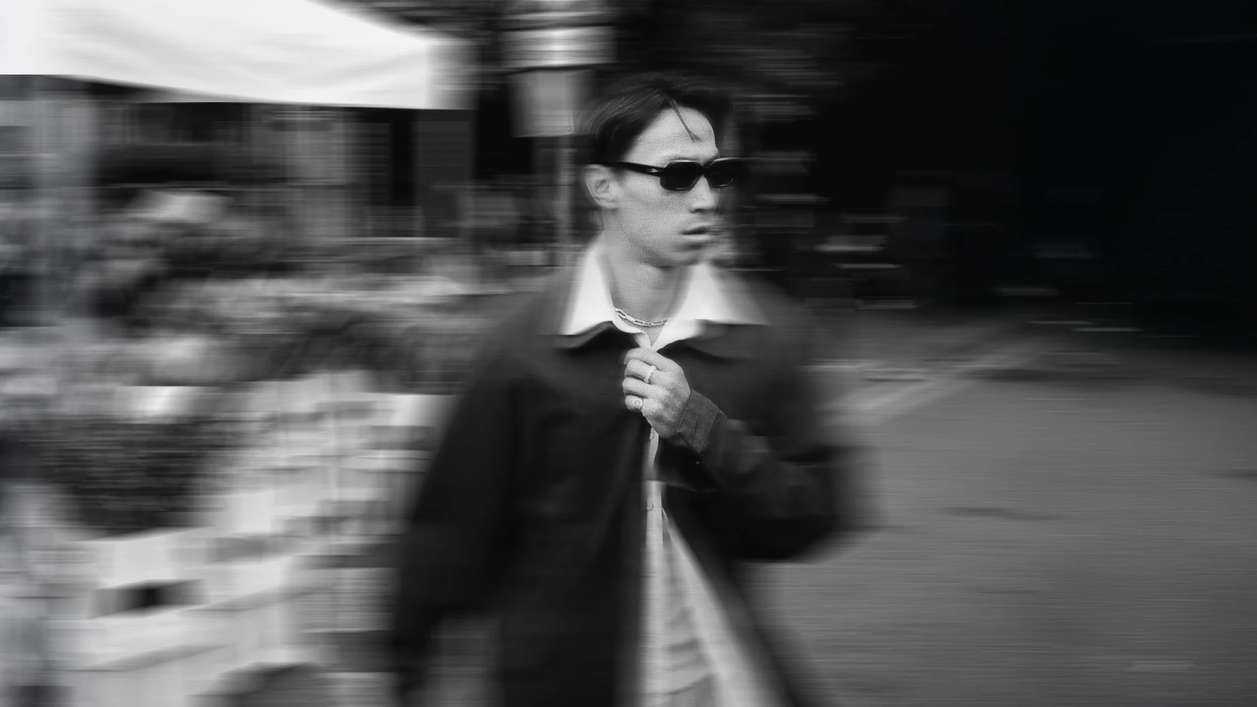 A person wearing sunglasses, a chain necklace, a collared shirt, and a jacket walking outdoors with motion blur in the background.