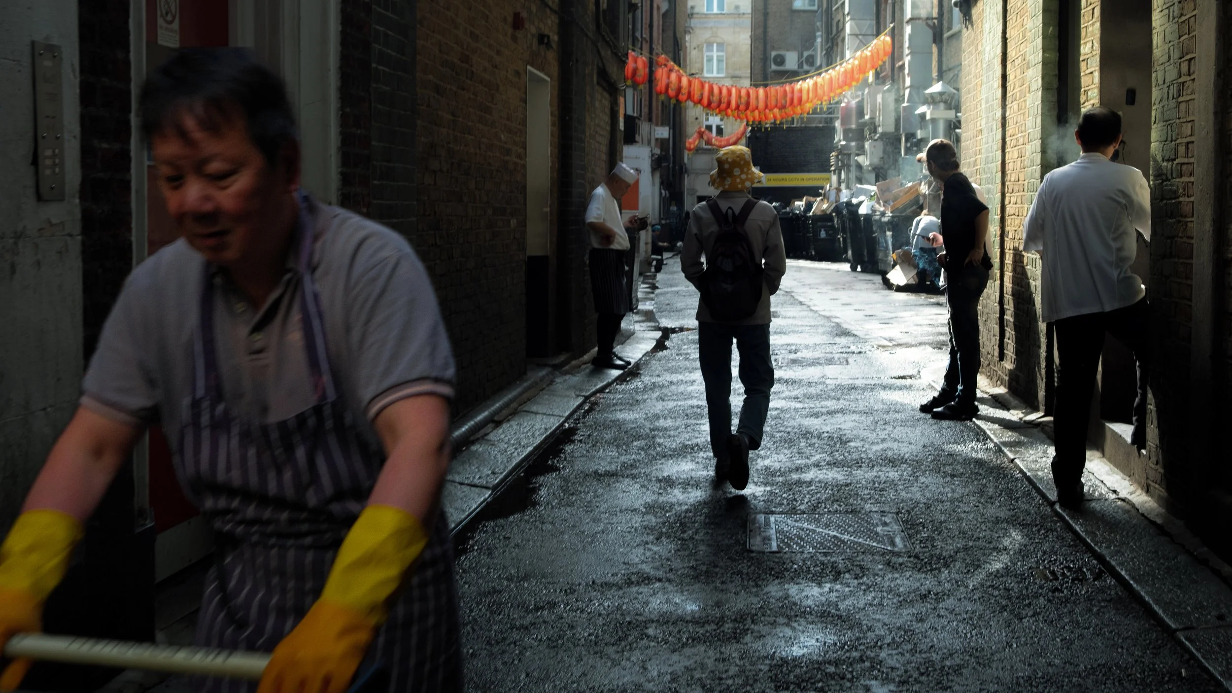An alleyway with several people, including a woman in a yellow hat and backpack, and a worker in yellow gloves cleaning the wet street, with hanging orange lanterns visible.