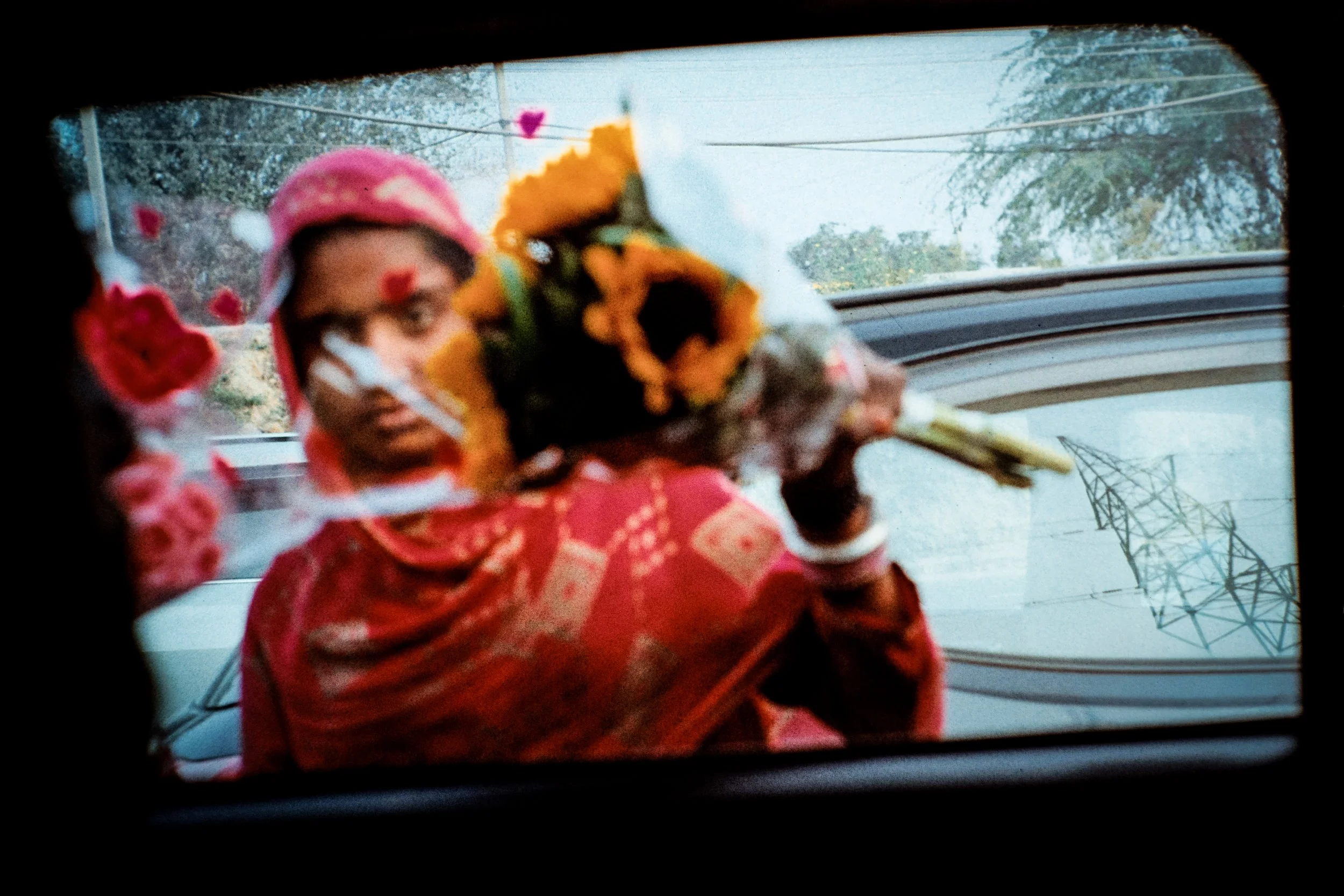 A woman dressed in traditional Indian attire holding a bouquet of sunflowers, seen through a car window with windshield wipers and power lines in the background.