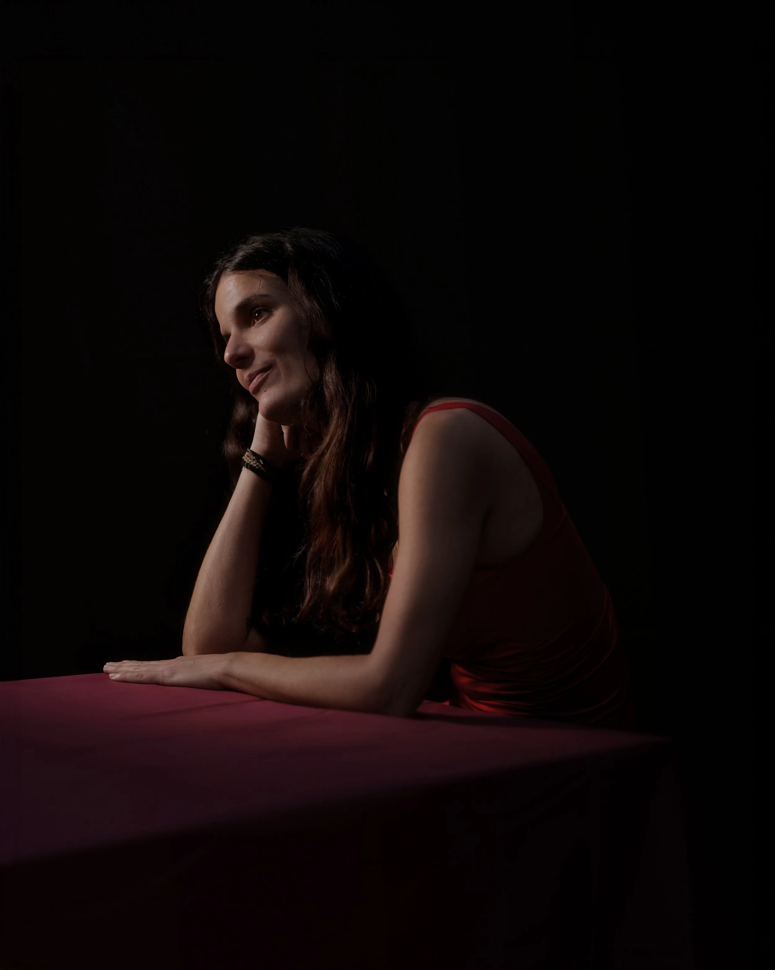 A woman with long dark hair sitting at a table with a red tablecloth, resting her chin on her hand, smiling slightly against a dark background.