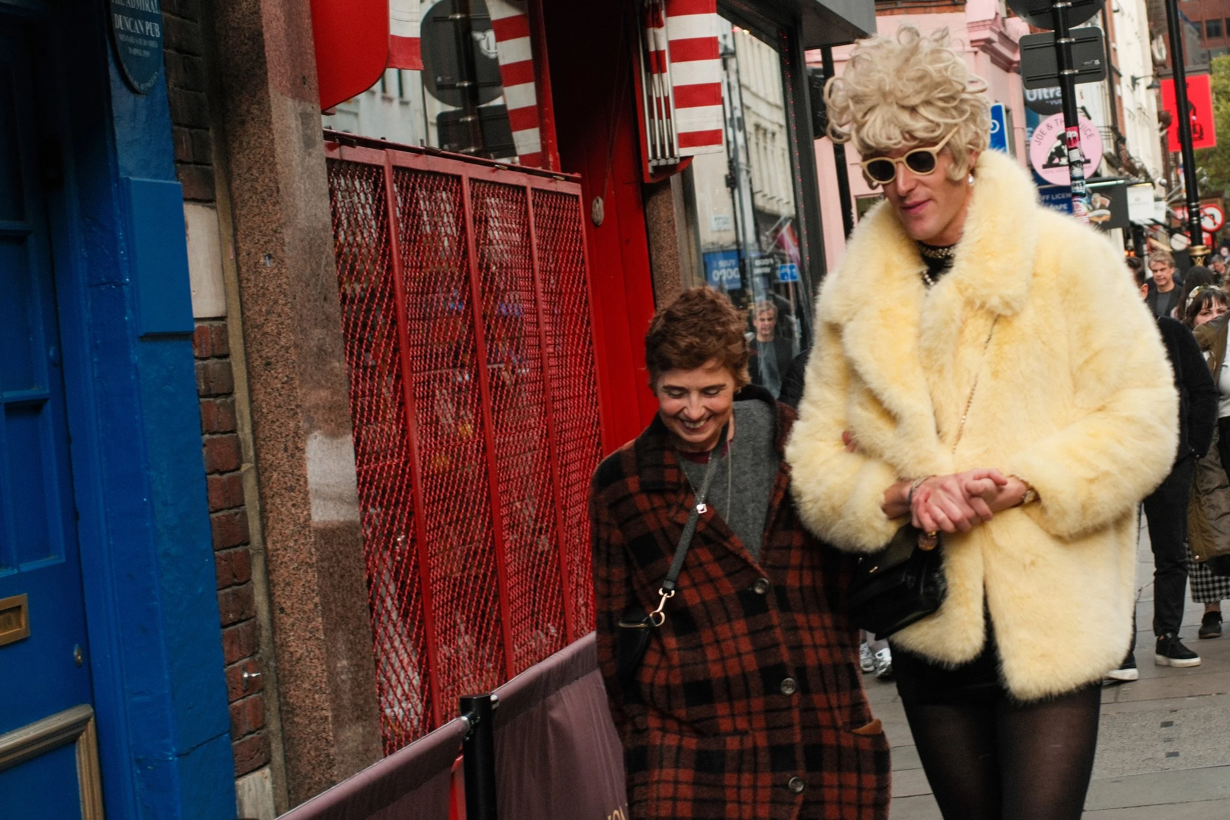 Two women smiling, walking together on a city sidewalk, one wearing a yellow faux fur coat and sunglasses, the other wearing a brown plaid coat, surrounded by city buildings and pedestrians.