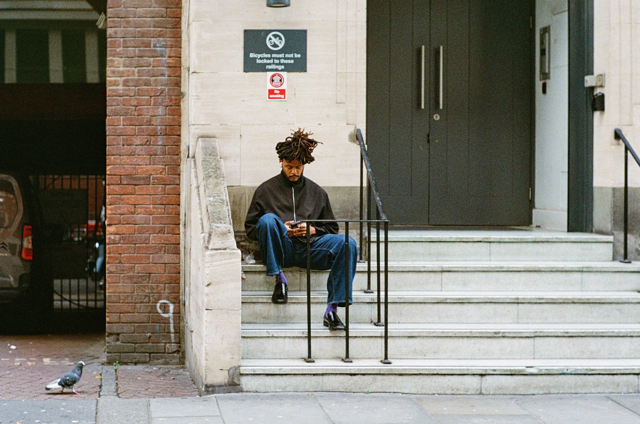 A man with dreadlocks seated on steps outside a building, looking at his phone, with a pigeon nearby and a sign indicating bicycles must not be locked to railings.