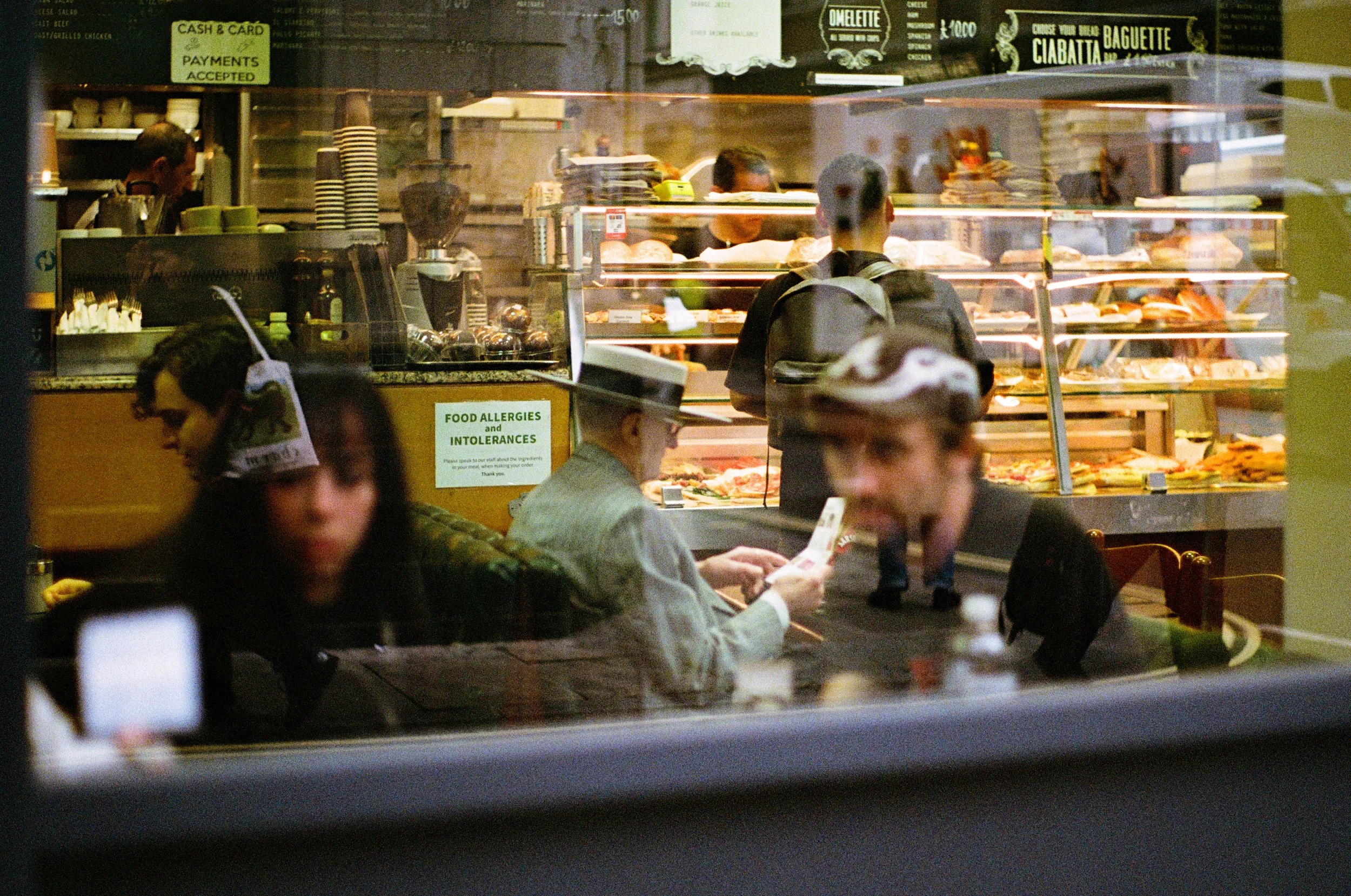 View through a restaurant window showing customers waiting and ordering, with a counter displaying baked goods and menu signs hanging above.