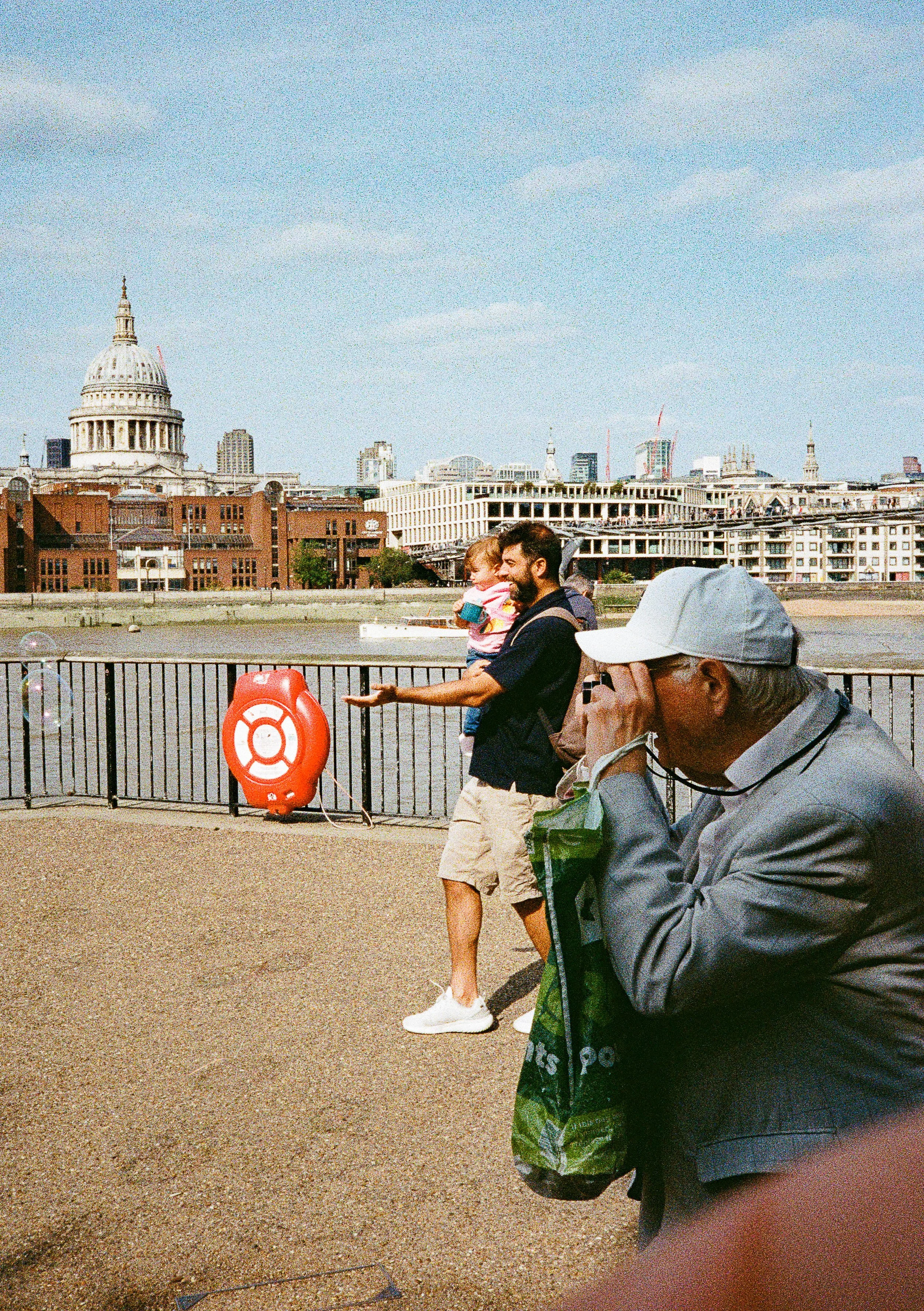 A man holding a young girl in his arms, standing near a railing with the London skyline, including the Shard and St. Paul's Cathedral in the background. An elderly man wearing a cap and glasses is looking at his phone near the right side of the image