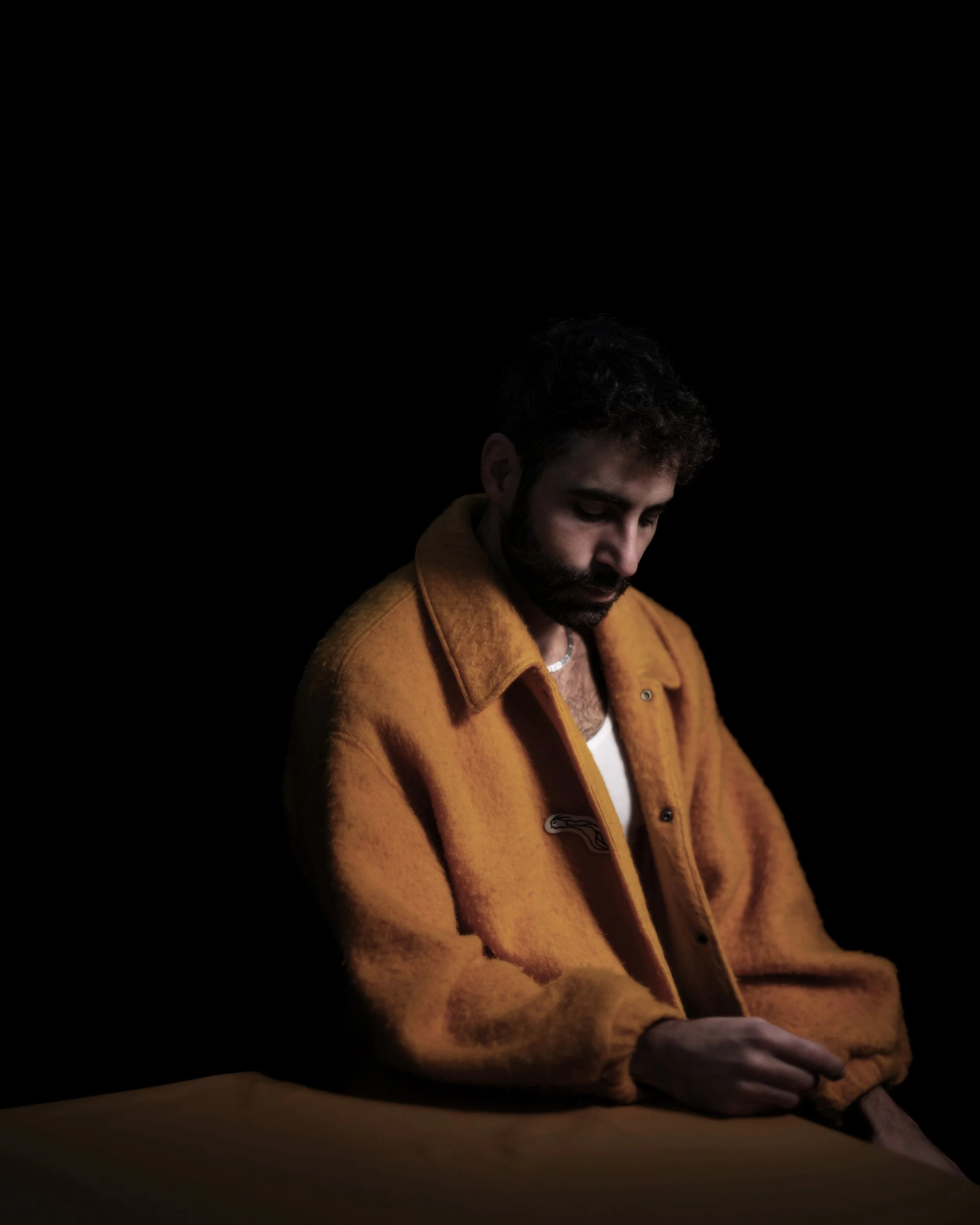 A man with a beard and dark curly hair wearing a brown jacket sitting at a table with a dark background.