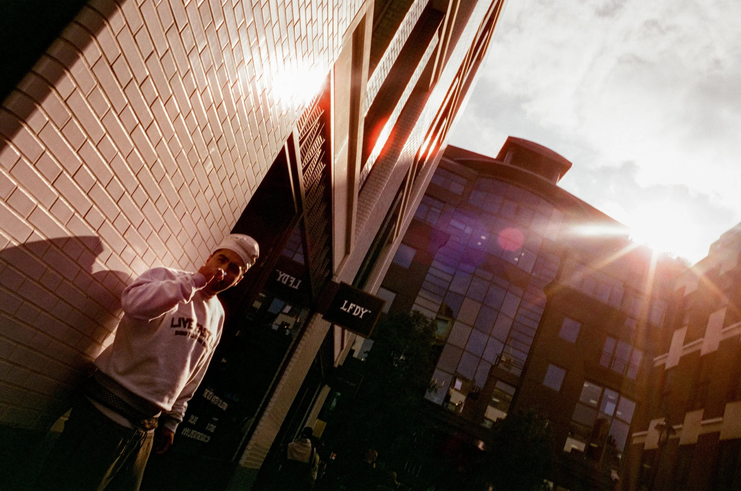 Person standing against a white brick wall in an urban area with tall buildings and bright sun flare in the sky.