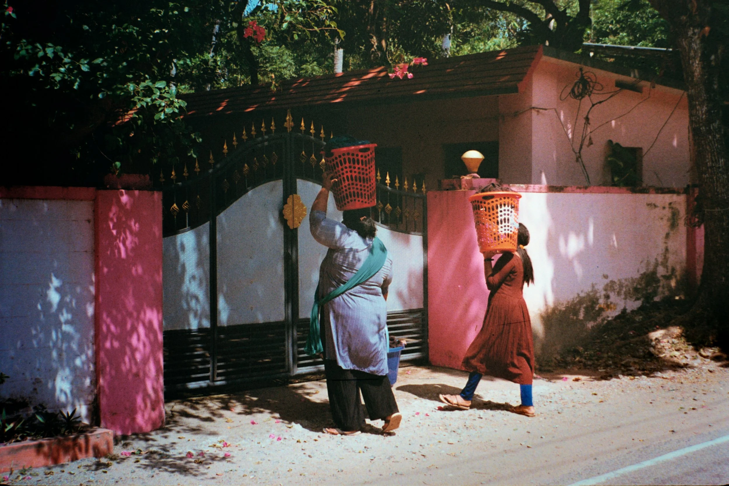 Two women walking along a street with baskets on their heads near a house with a pink and white wall and black gate, surrounded by trees and shaded areas.