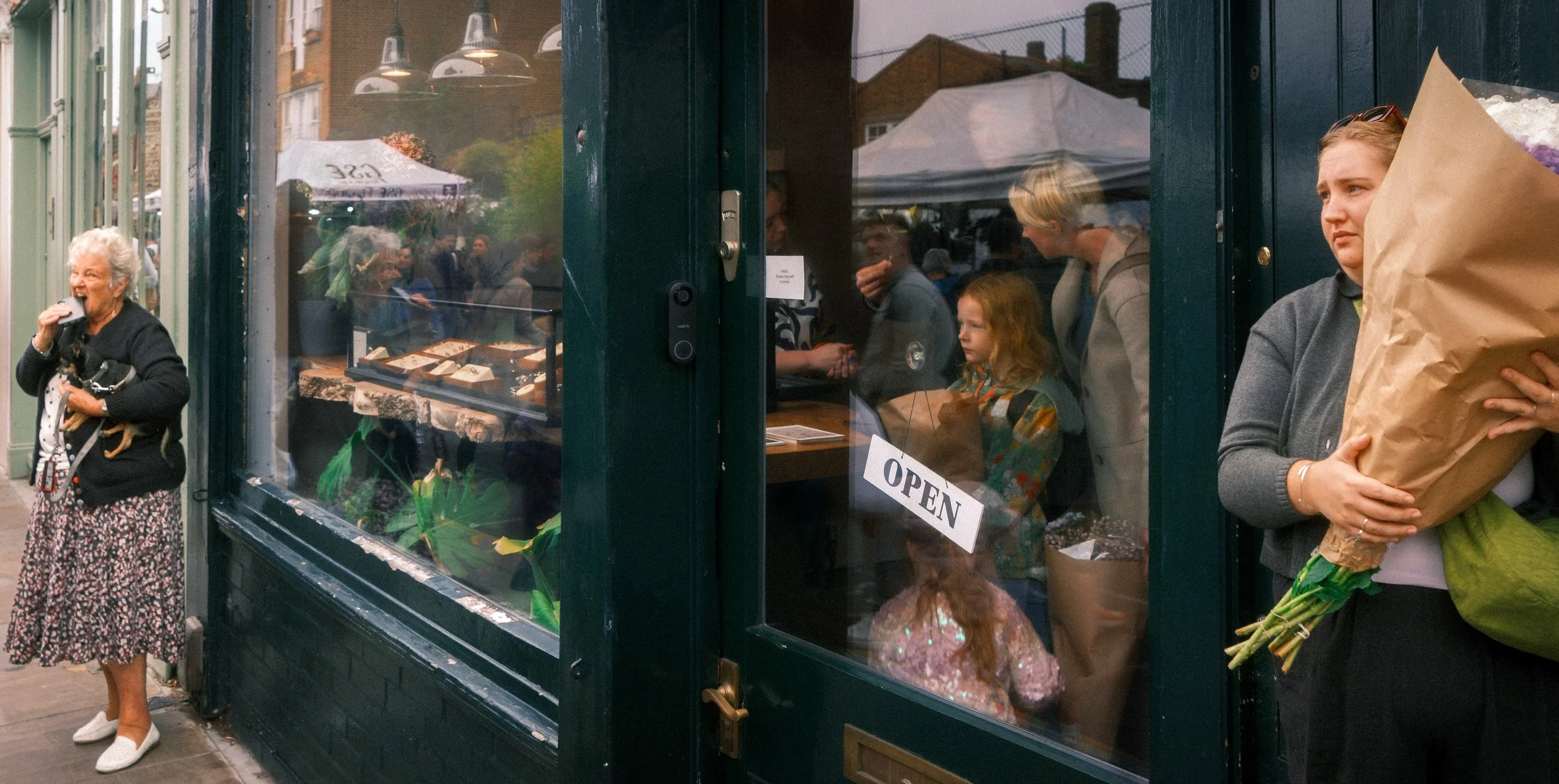 A woman with gray hair, wearing a black cardigan and a floral skirt, stands outside a storefront holding a small dog and eating a sandwich. Inside the store, visible through the window, are several people including a woman with or blonde hair and a c