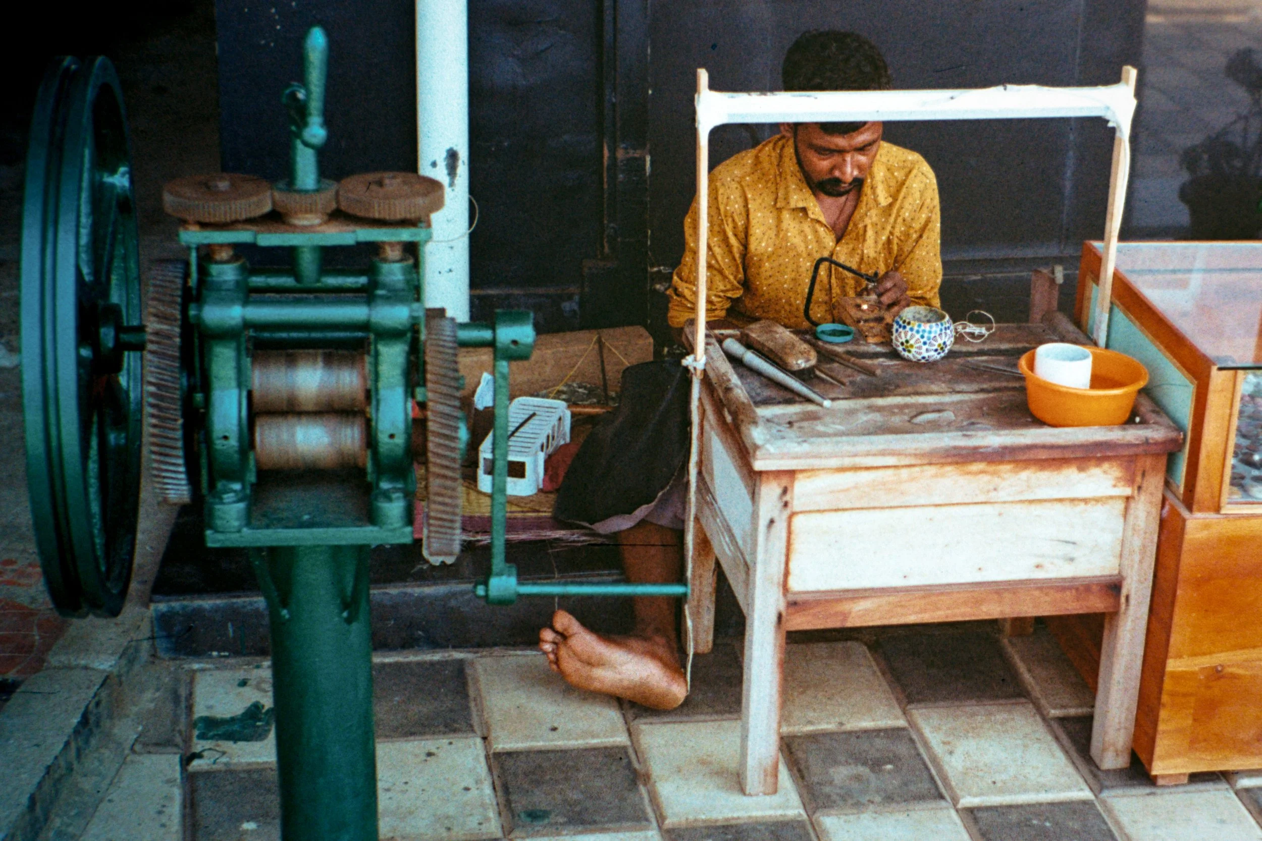 A man working at a small wooden table with a glass of water, a bowl, and tools, while sitting beside a vintage green money-making machine, and his foot is visible underneath the table.