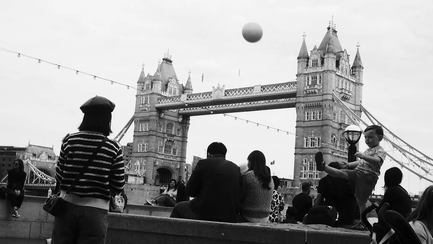 People sitting and standing near Tower Bridge in London, with a child playing and a woman in a striped shirt in the foreground, black and white photo.