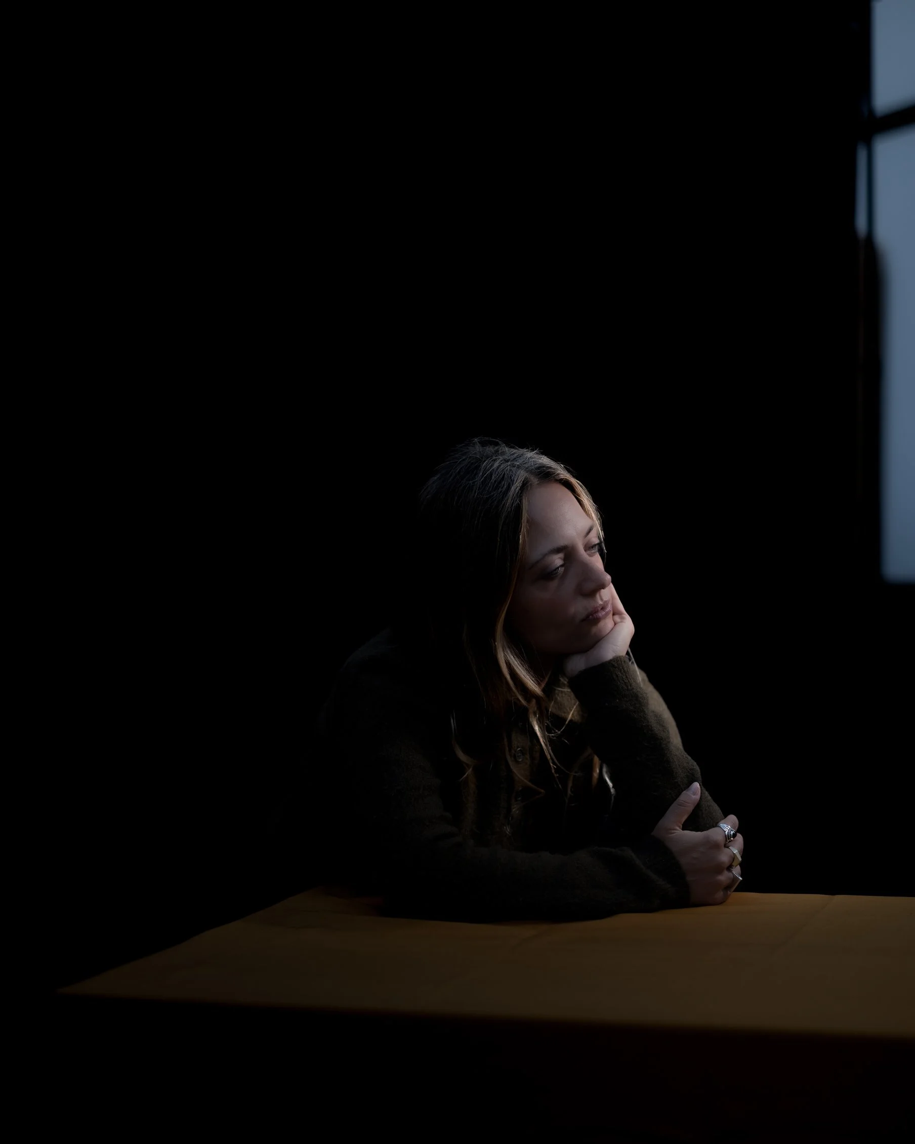 A woman with brown hair sitting at a table, resting her head on her hand, looking thoughtful, with light coming from the window on the right side of the image.