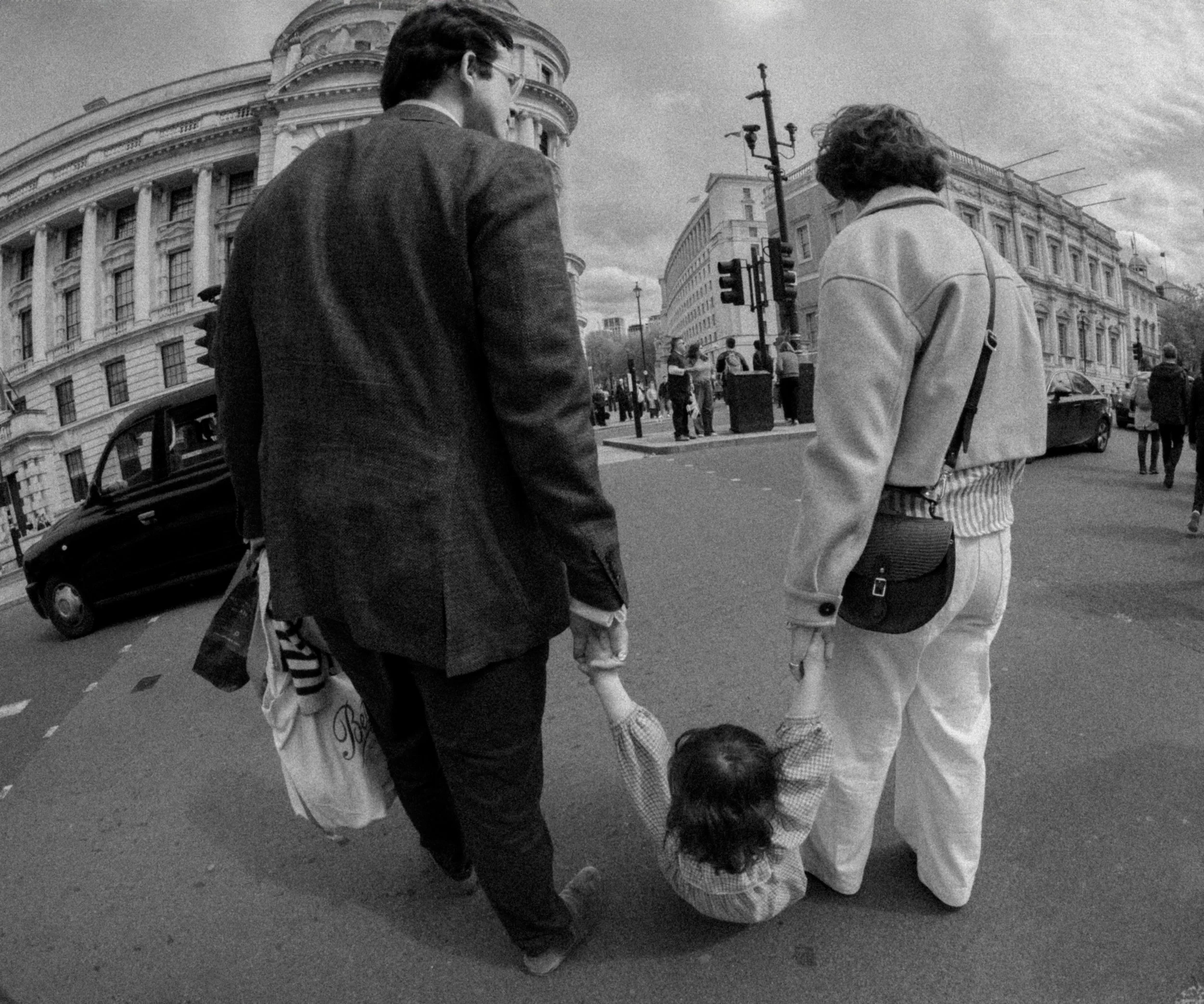 Black and white photo of two adults walking with a small child holding their hands on a city street, with buildings and other pedestrians in the background.