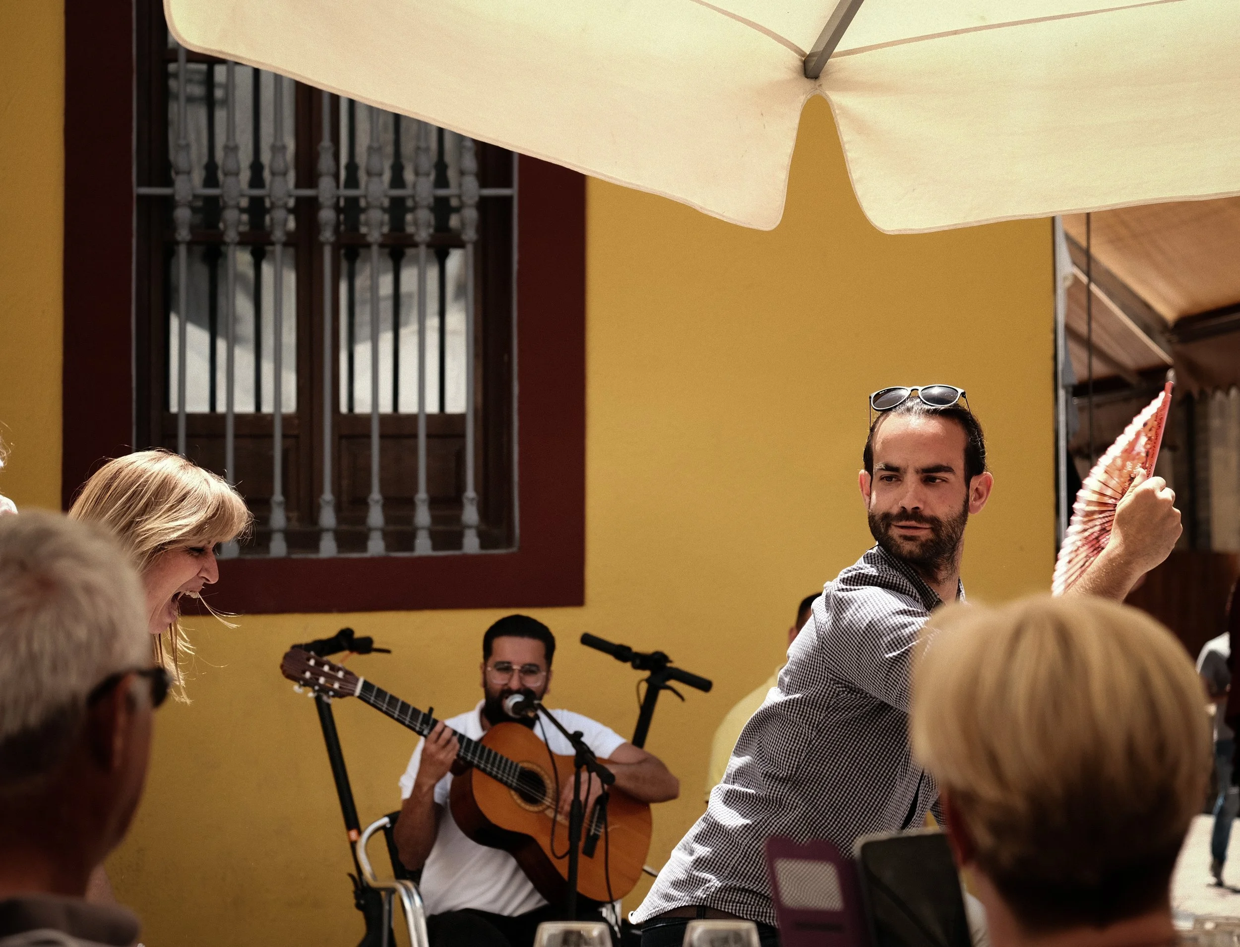 A man with sunglasses on his head and a fan in his hand poses at an outdoor gathering. In the background, a musician plays a guitar and sings into a microphone while several people listen.