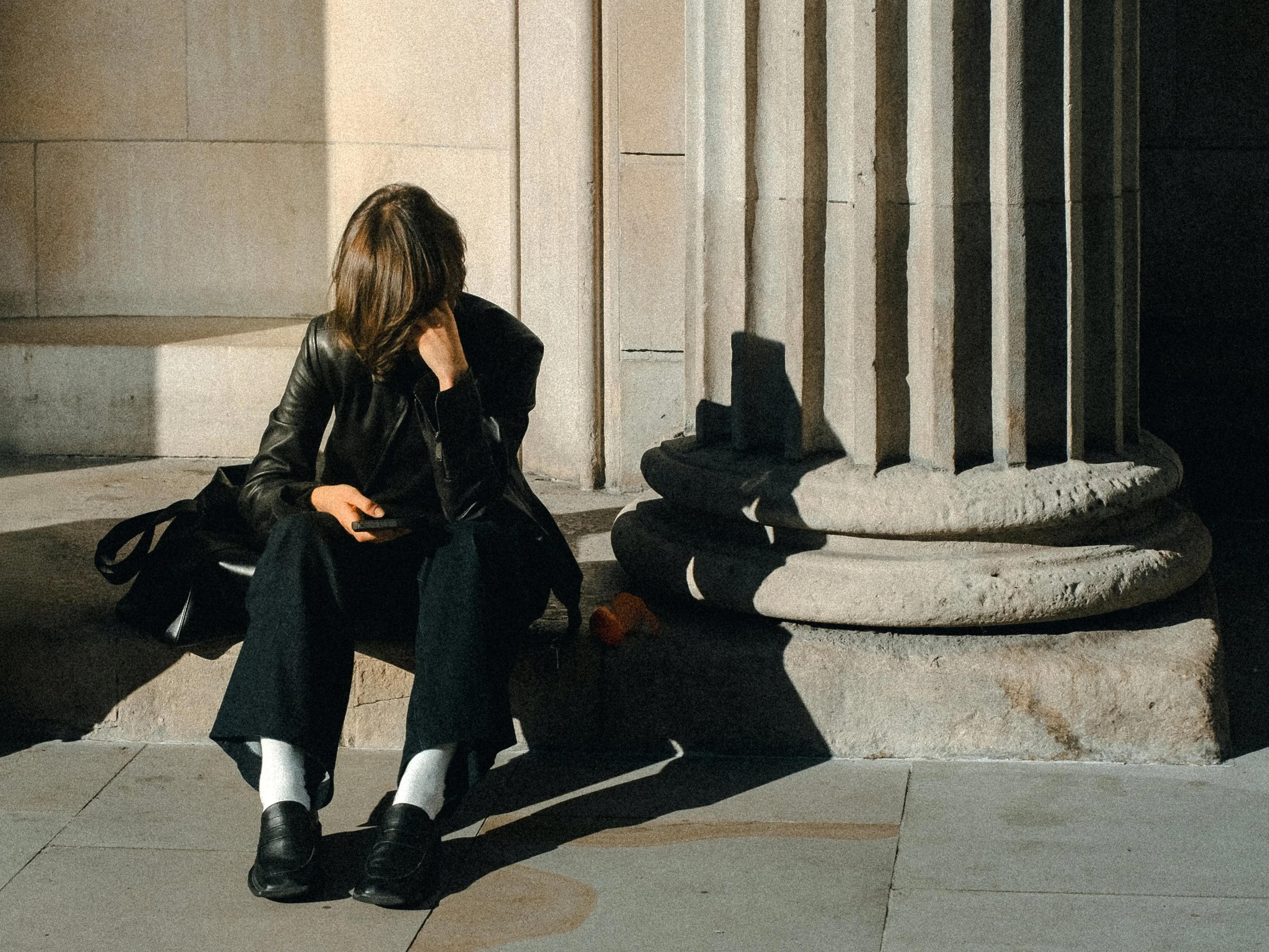 Woman sitting on the ground next to a large stone column, looking at her phone, with her face partially obscured by her hair and hand, wearing a black leather jacket, black pants, white socks, and black shoes.