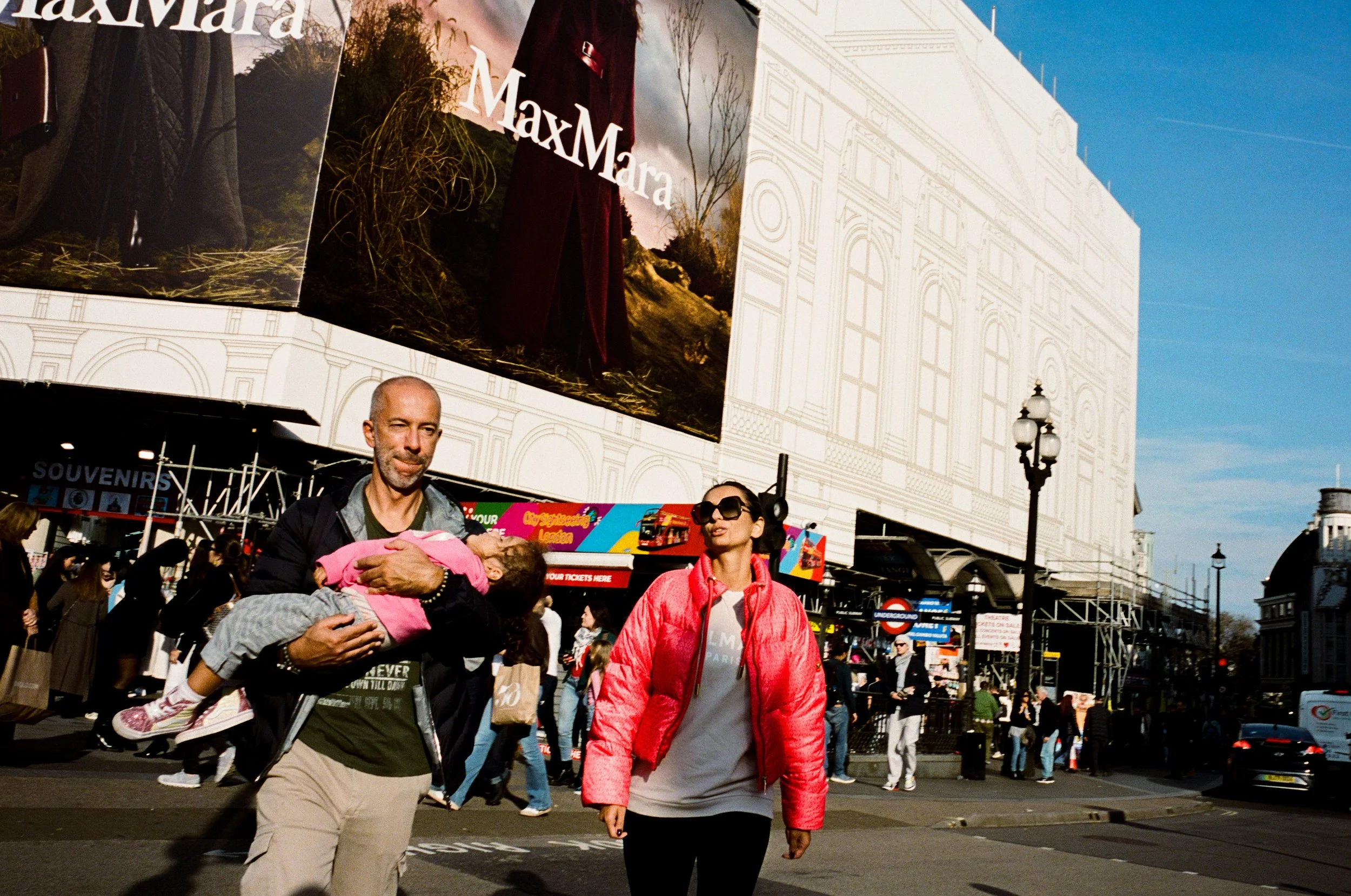 A man holding a sleeping child, a woman walking on a busy street in front of a large advertising billboard for MaxMara, with many pedestrians and city buildings in the background.