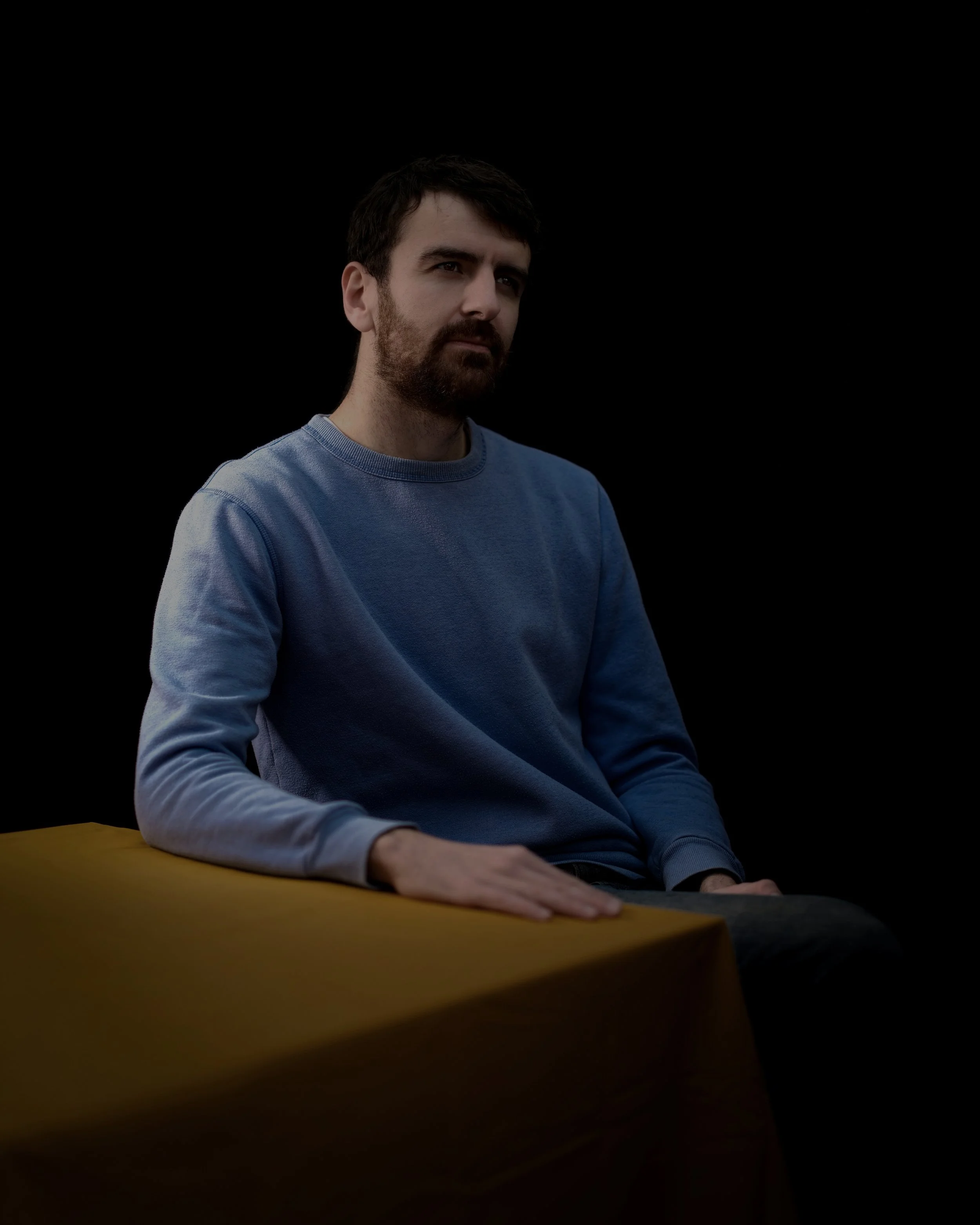 A man with dark hair and a beard, wearing a blue sweatshirt, sitting at a table with a yellow cloth against a black background.
