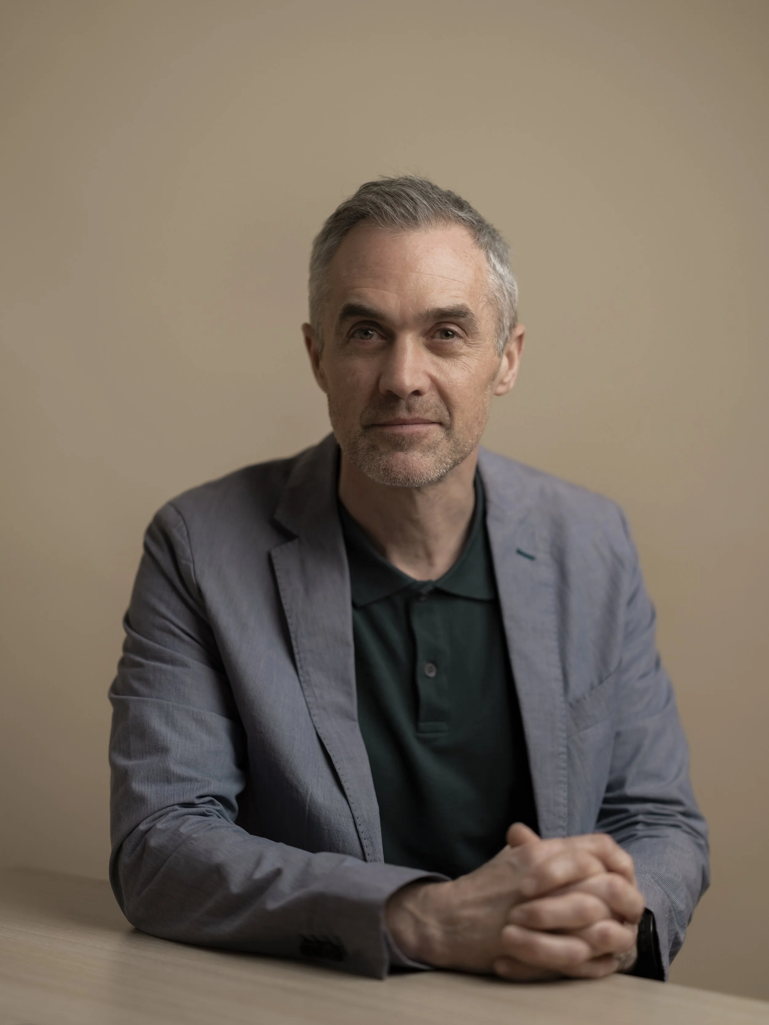 A middle-aged man with gray hair and a slight beard sitting at a table against a beige background, wearing a gray blazer and a dark green polo shirt, looking at the camera.