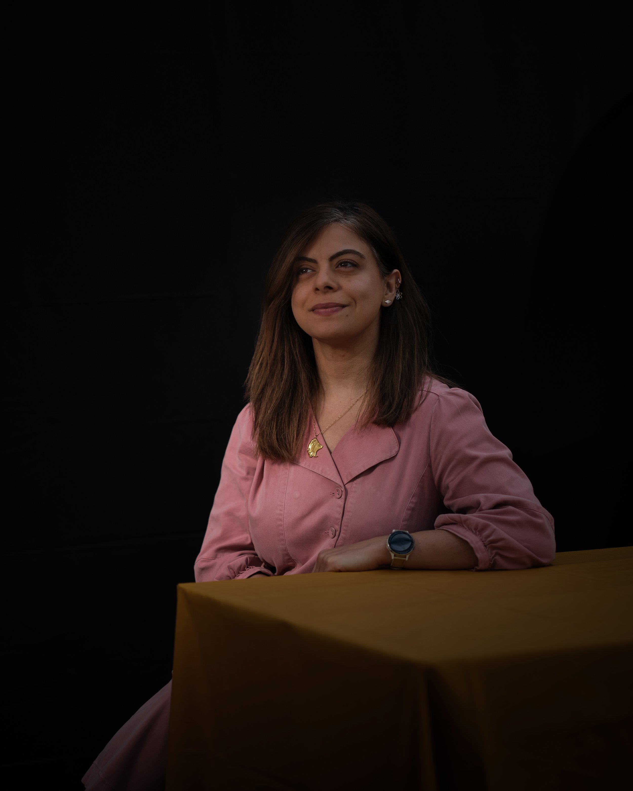 A woman with shoulder-length brown hair sitting at a table, wearing a pink shirt, a gold necklace, and a watch, with a dark background.