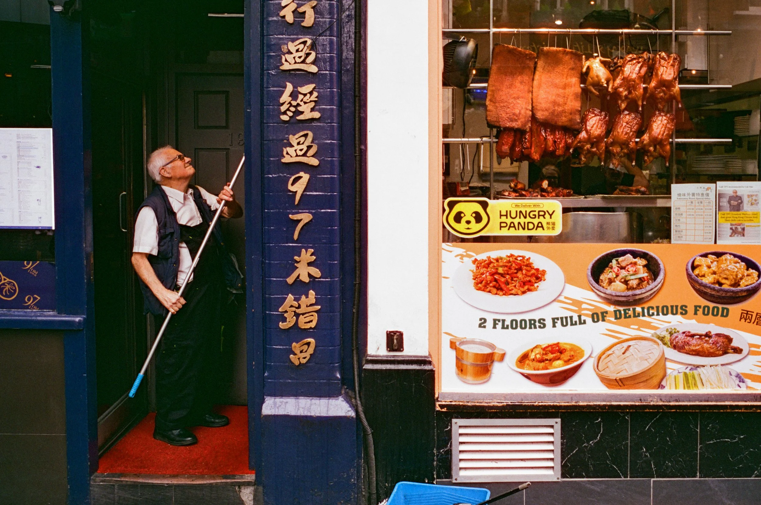 An older man with glasses, white hair, wearing a white shirt and black vest, holding a broom, standing at the entrance of a restaurant with Chinese characters on a vertical sign. The restaurant has roasted meats hanging inside and a colorful sign adv