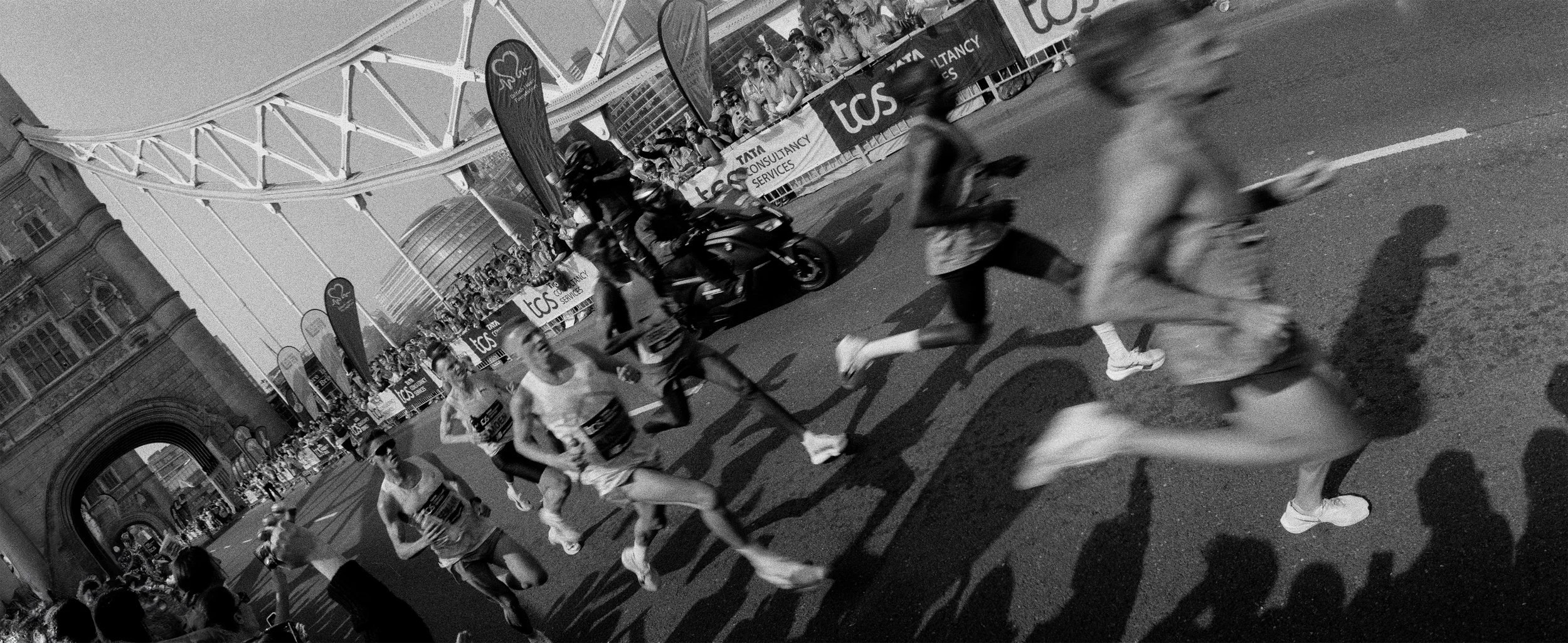 Black and white photo of a night marathon race with runners running on a city street, some on motorbikes, and spectators behind barriers.
