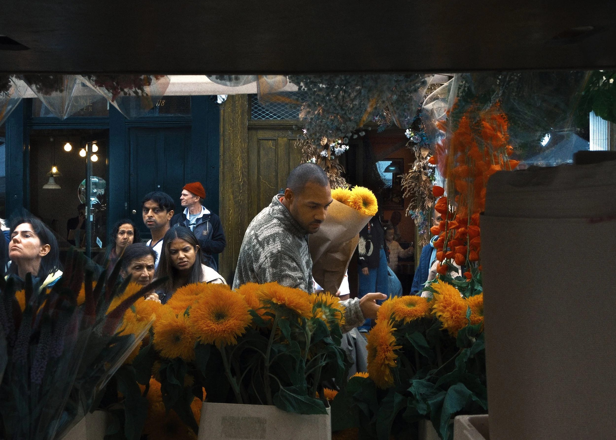 People shopping at a flower market, focusing on a man with a bouquet of yellow flowers, surrounded by numerous other people and colorful flowers.