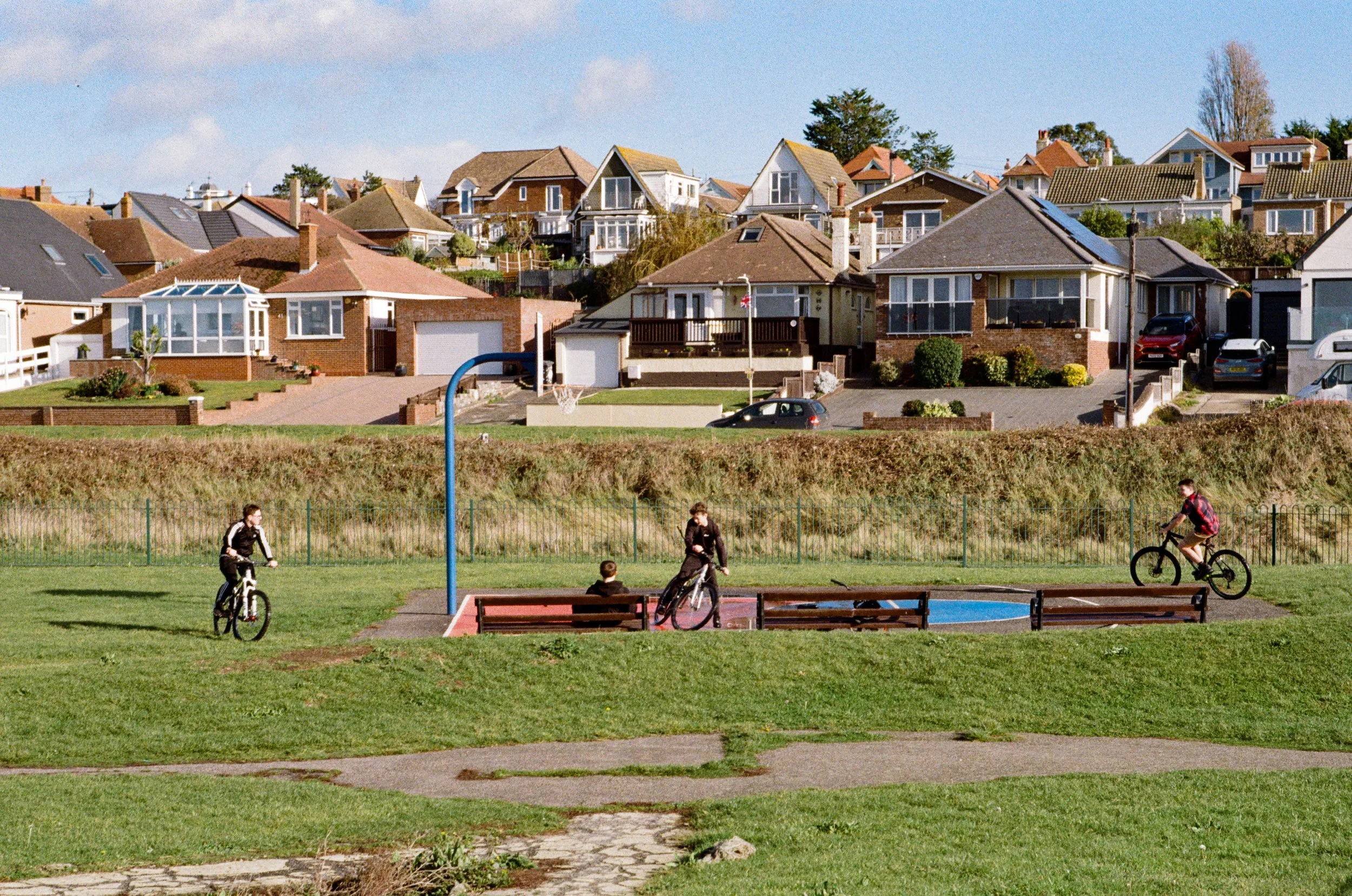 Children riding bikes and sitting on benches in a park near a playground with a basketball hoop, with residential houses on a hillside in the background.