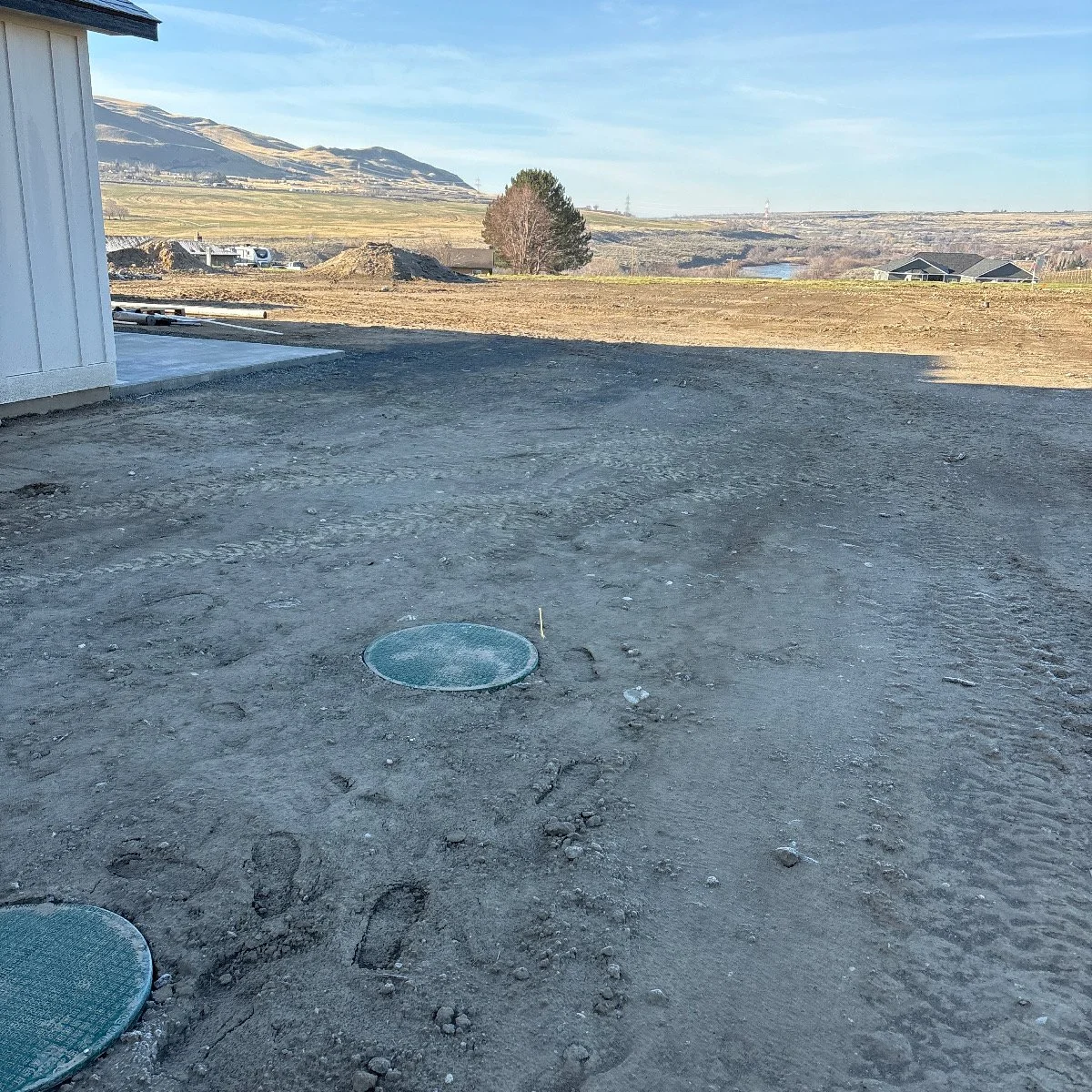 A dirt area with two visible manhole covers, part of a building on the left, and a scenic landscape with hills, trees, and houses in the background under a clear sky.