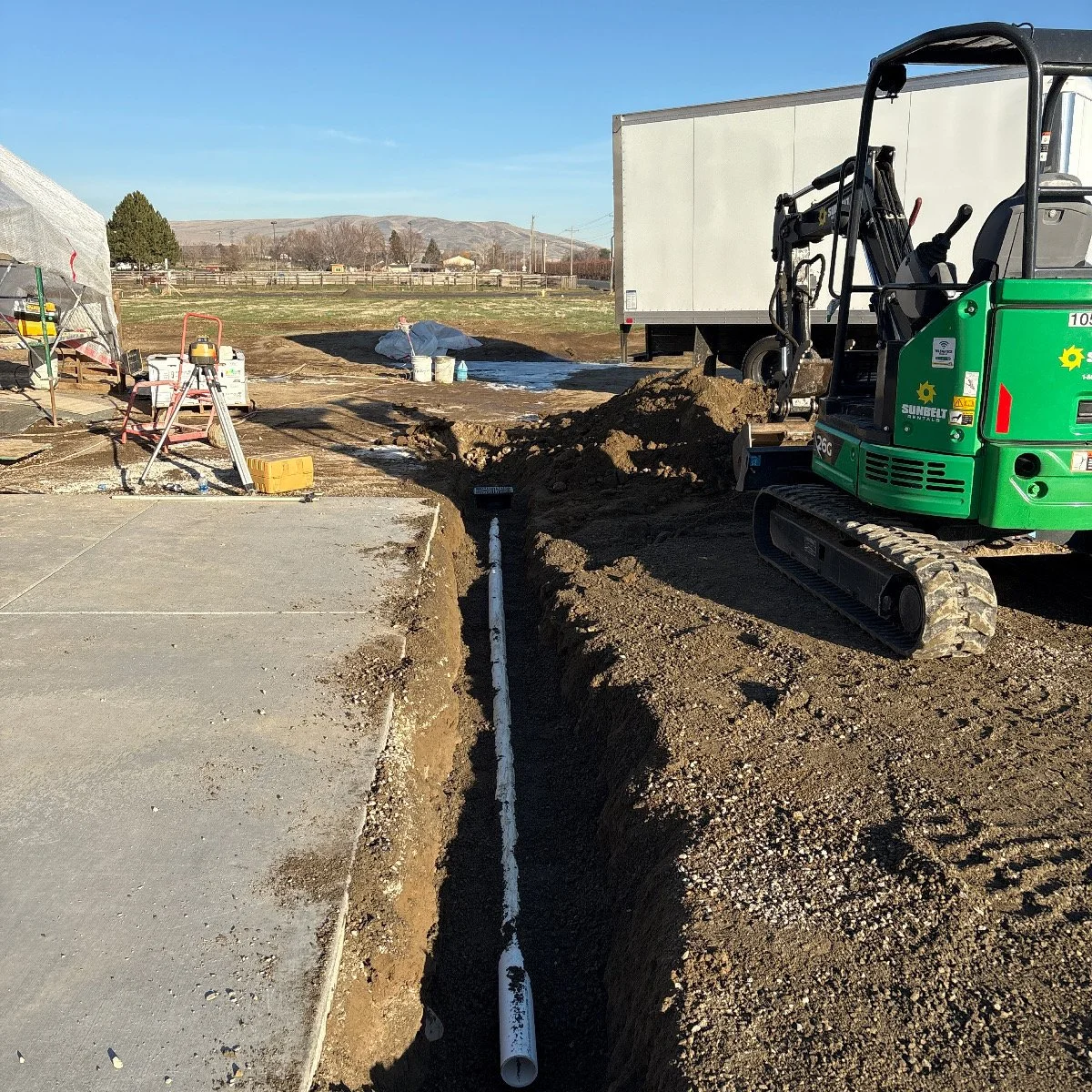 Construction site with a small excavator digging a trench and a pipe run through the trench. There is construction equipment and materials around, and an overcast sky.