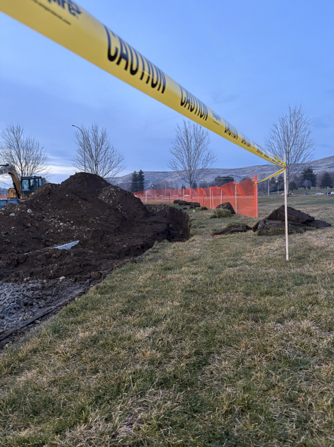 Construction site with an open trench, yellow caution tape, a pile of dirt, orange safety fence, and leafless trees in the background.