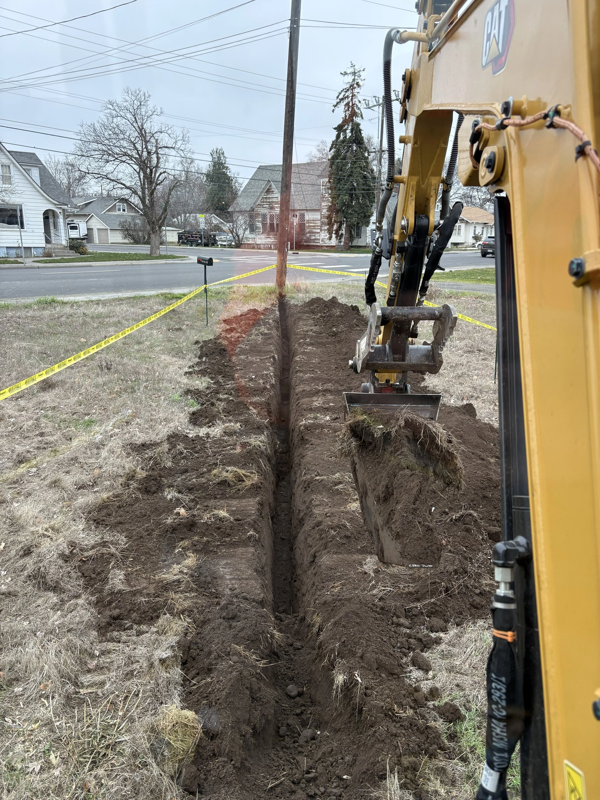 A yellow excavator digging a trench in a grassy area near a residential street, with construction tape cordoning off the area.