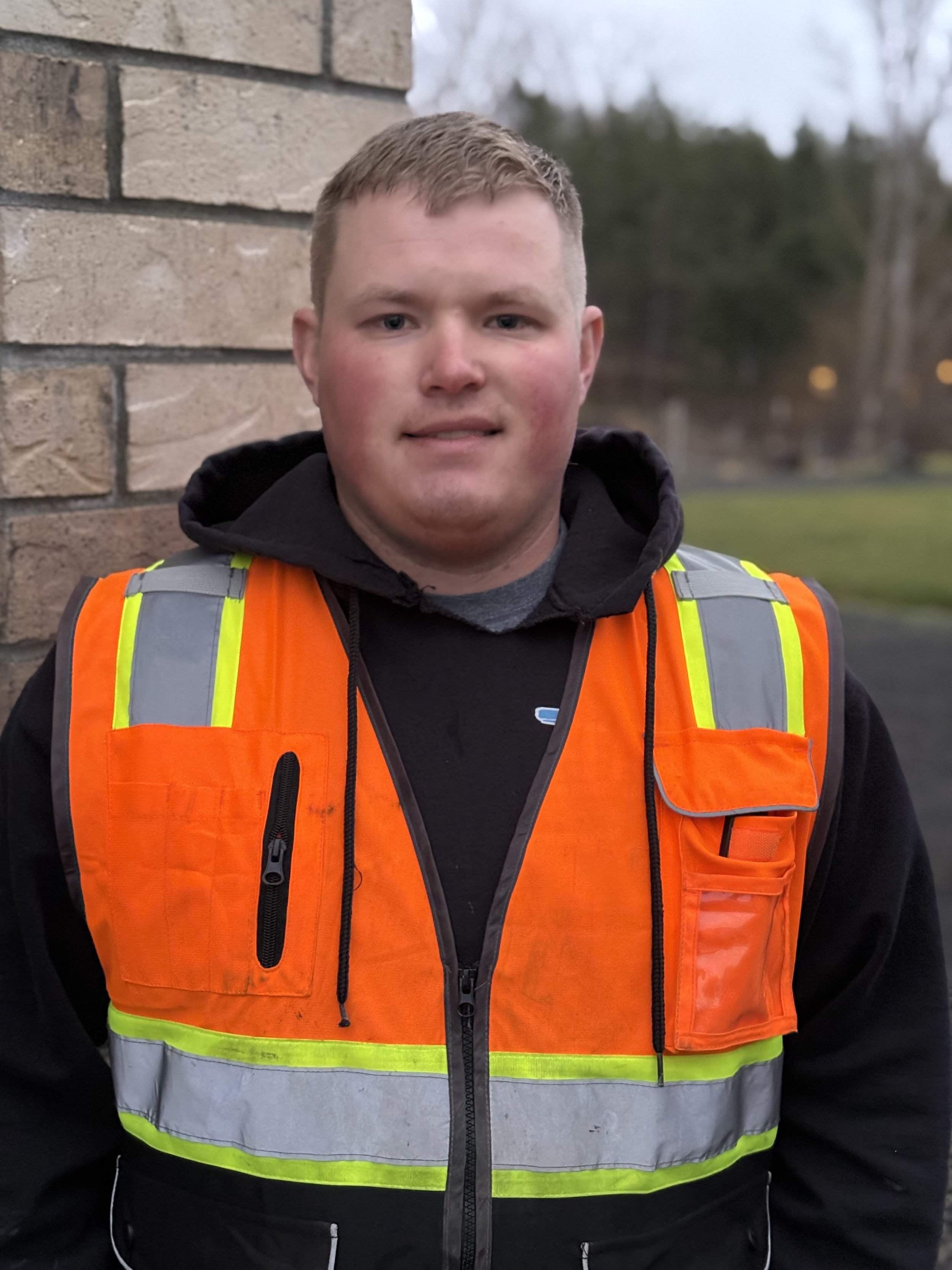 A man wearing an orange safety vest lined with reflective silver stripes, standing outdoors against a brick wall with a blurred background of trees and open fields.