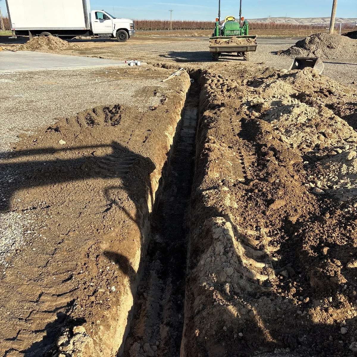 A construction site showing a deep trench in the ground with piles of dirt on each side, a small green tractor in the background, a white utility truck, and various construction tools and materials nearby.