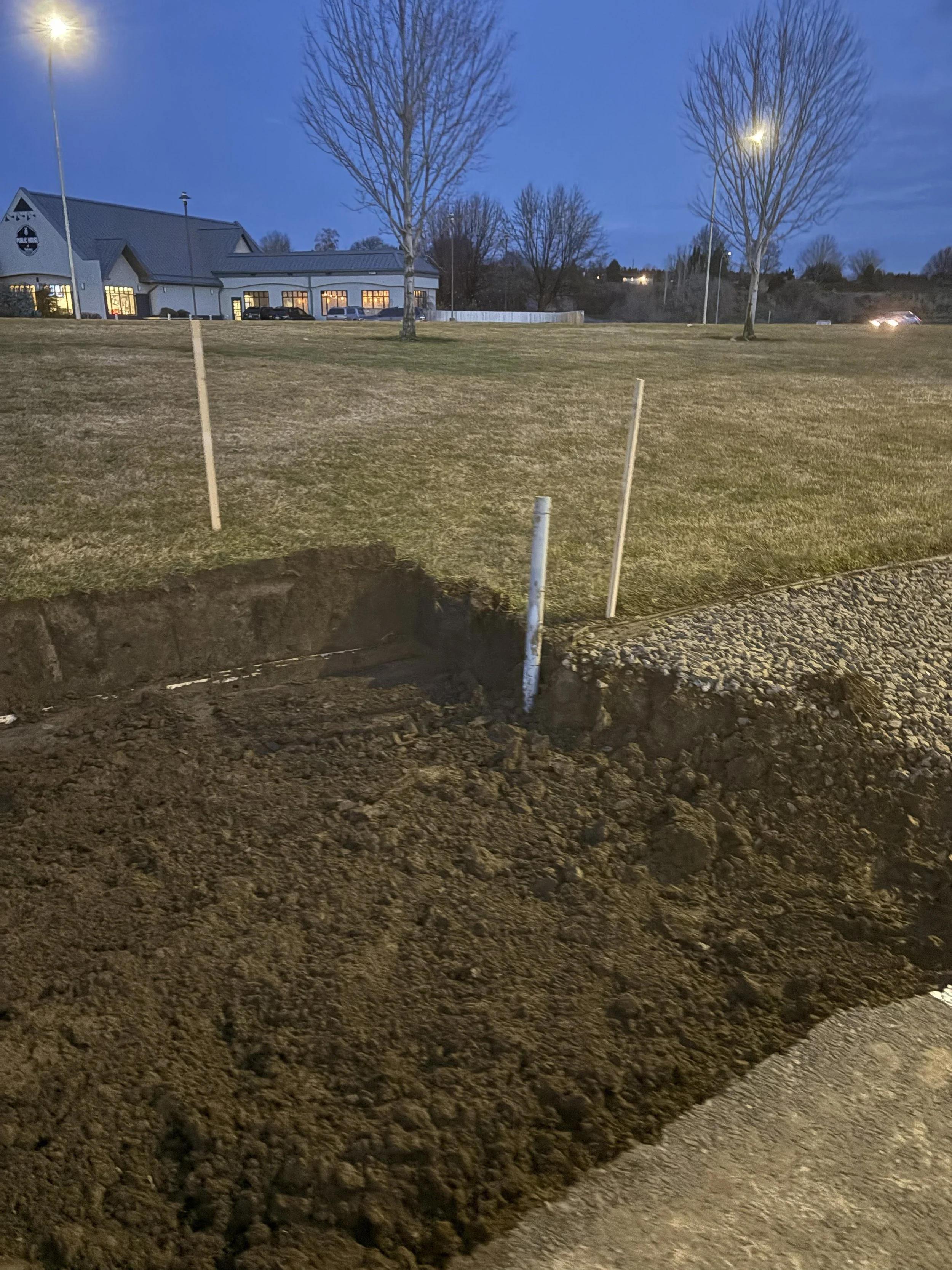 A construction site at dusk with a large hole in the ground, wooden stakes marking the area, and around the site is a grassy area with a commercial building and trees in the background.