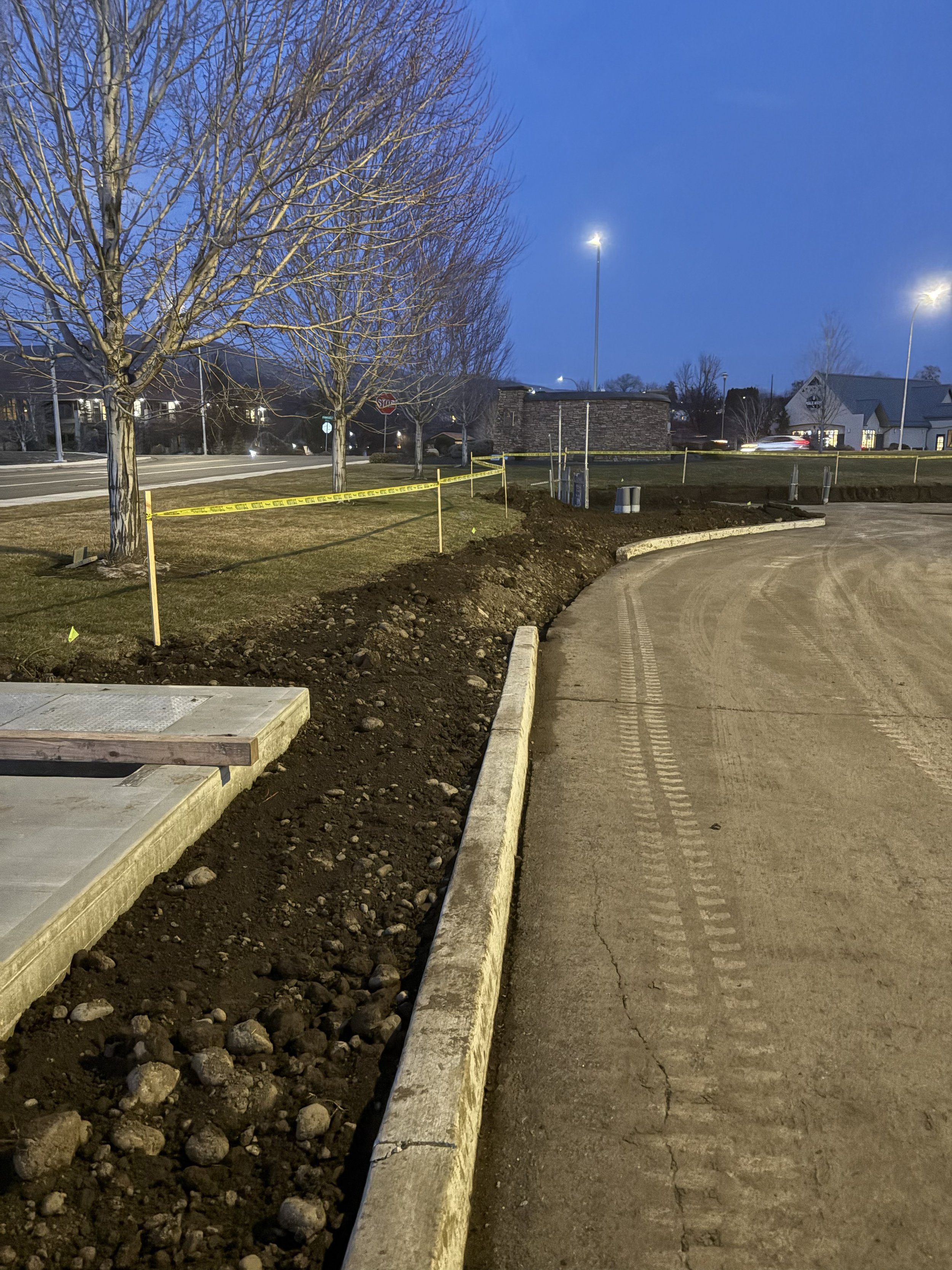 Construction site of a sidewalk or road extension at dusk with trees, caution tape, and a small brick building in the background