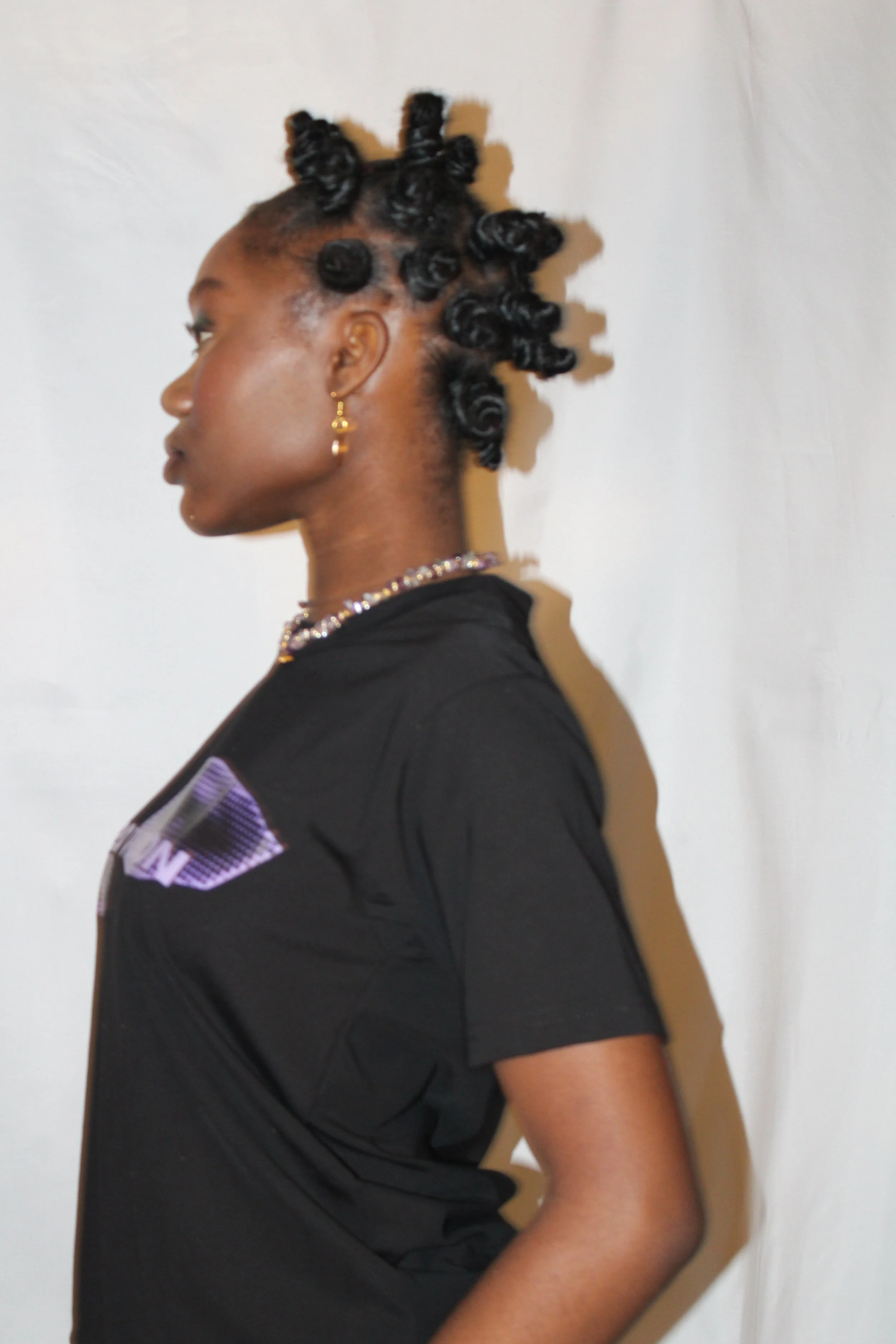 Side profile of a woman with styled bantu knots and gold hoop earrings, wearing a black T-shirt with a graphic design, standing against a plain white background.