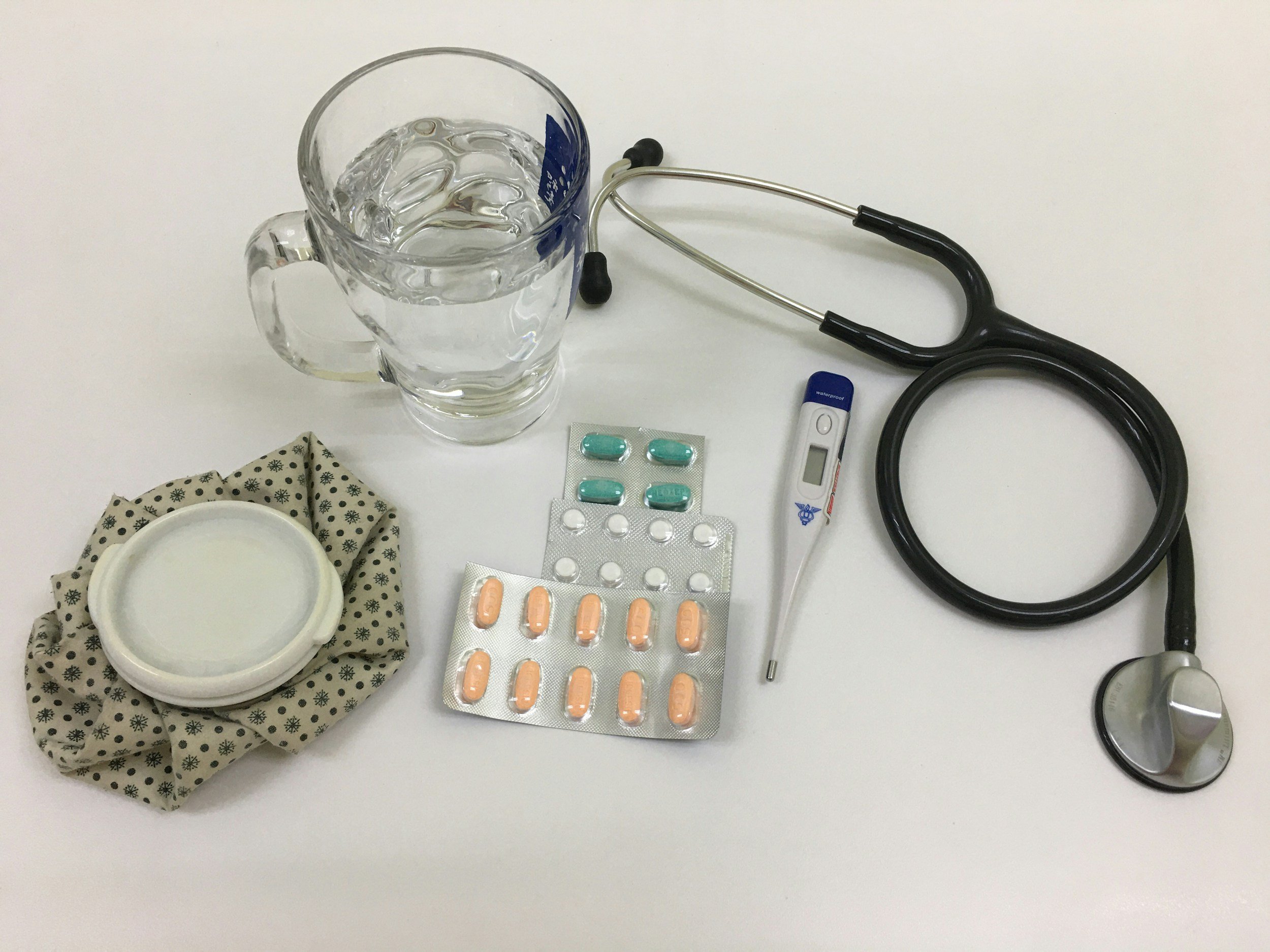 A glass of water, a stethoscope, a digital thermometer, a blister pack of capsules, a blister pack of tablets, a small container with ointment, and a cotton swab on a white surface.