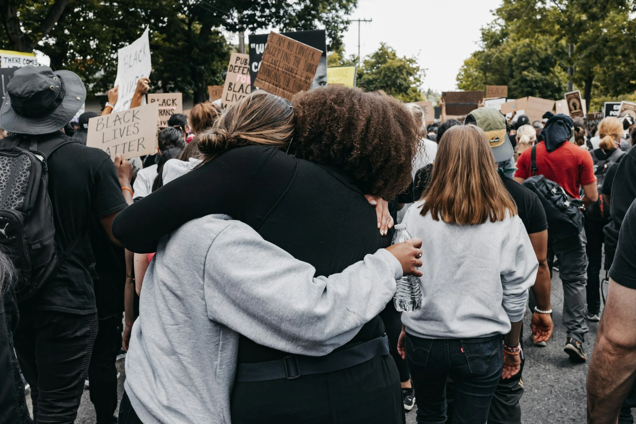 Two women hugging at a Black Lives Matter protest, surrounded by a crowd holding signs with messages supporting racial justice.