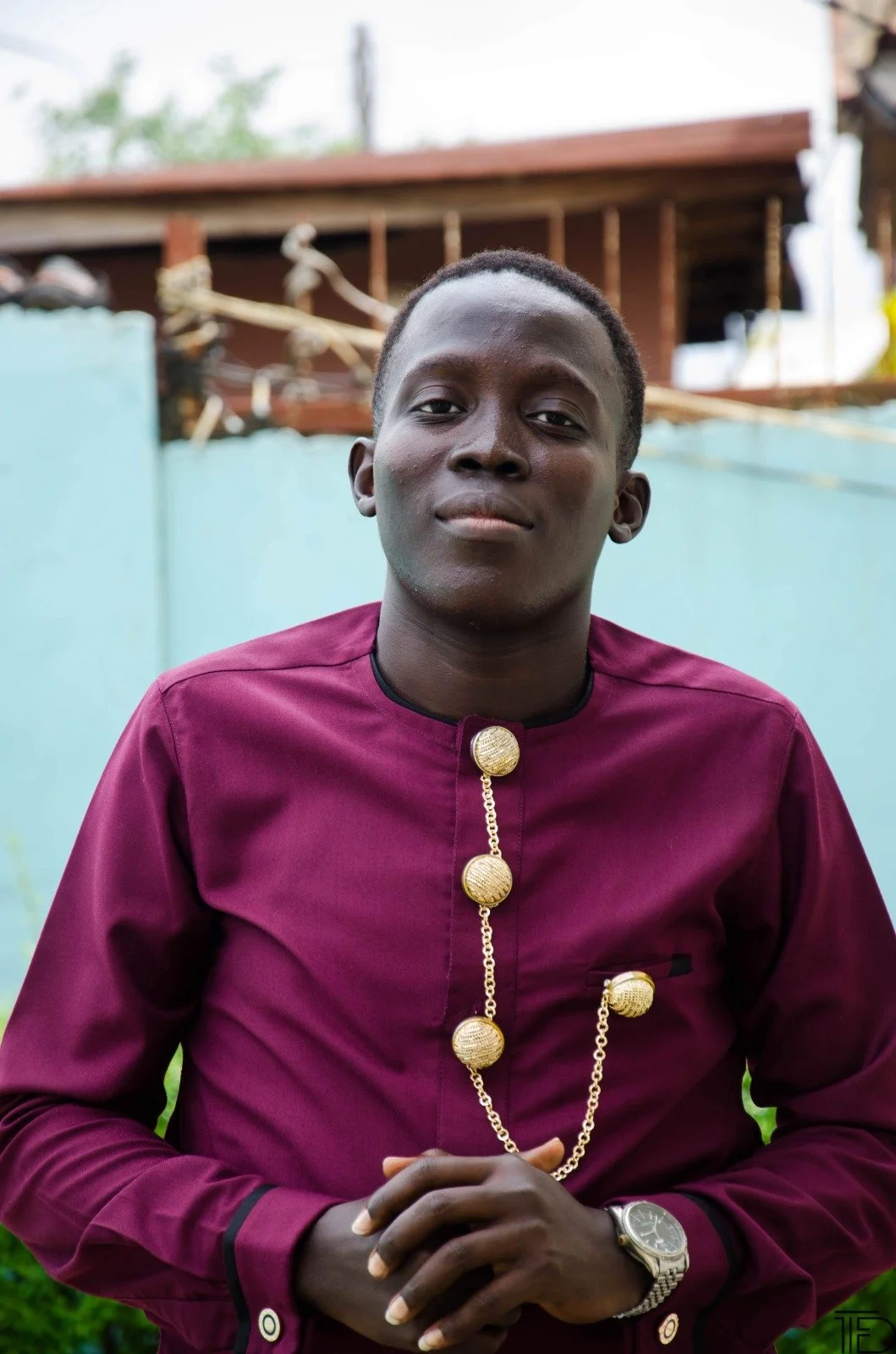 A young man wearing a maroon shirt with a gold chain necklace and a wristwatch, standing outdoors in front of a light blue wall.