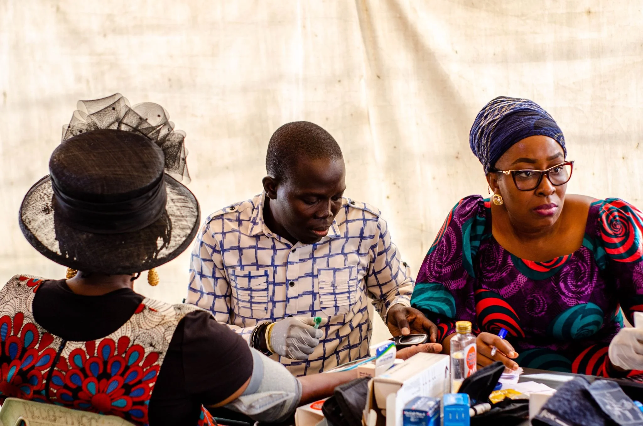 Two women and one man sitting at a table with medical supplies, with a plain beige background. The woman on the right is wearing glasses and a colorful headscarf, the man in the middle is taking notes, and the woman on the left is wearing a large black hat with a veil.