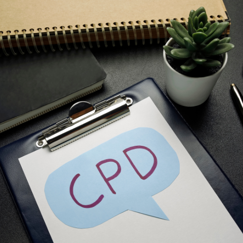 A clipboard with a paper showing the acronym "CPD" inside a blue speech bubble, a small potted succulent plant, a closed notebook, a spiral notebook, and a pen on a dark desk.